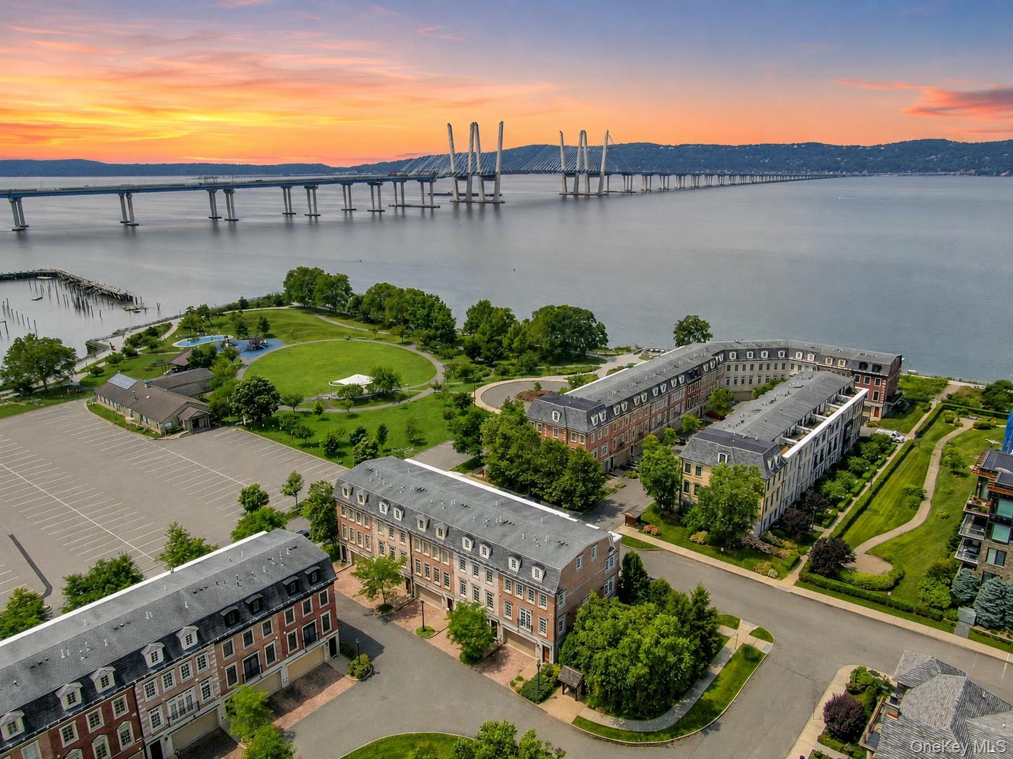 Aerial view of waterfront buildings, a landscaped park, and a cable-stayed bridge at sunset.