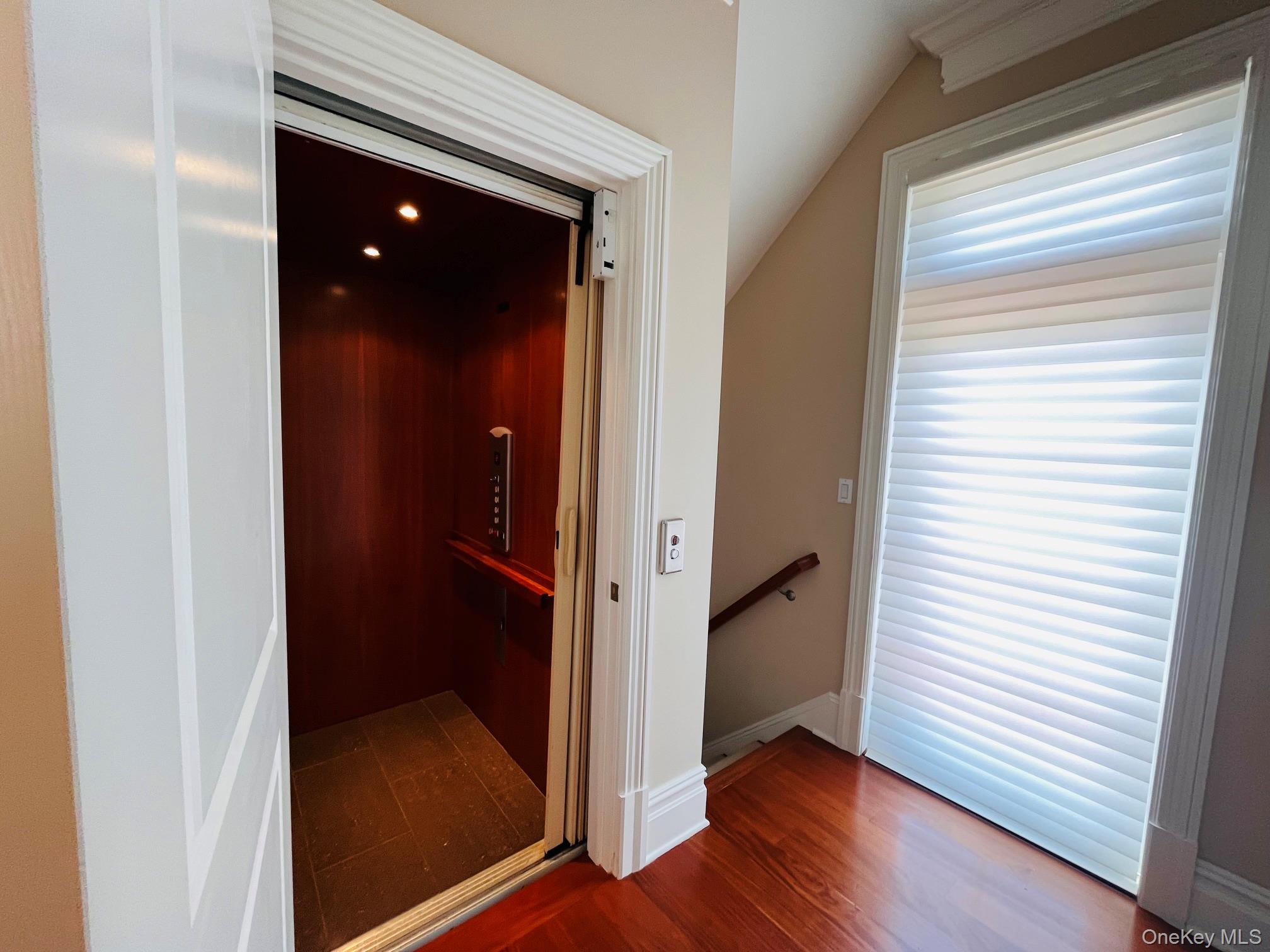Residential elevator beside a hardwood landing, staircase, and large window with white blinds.