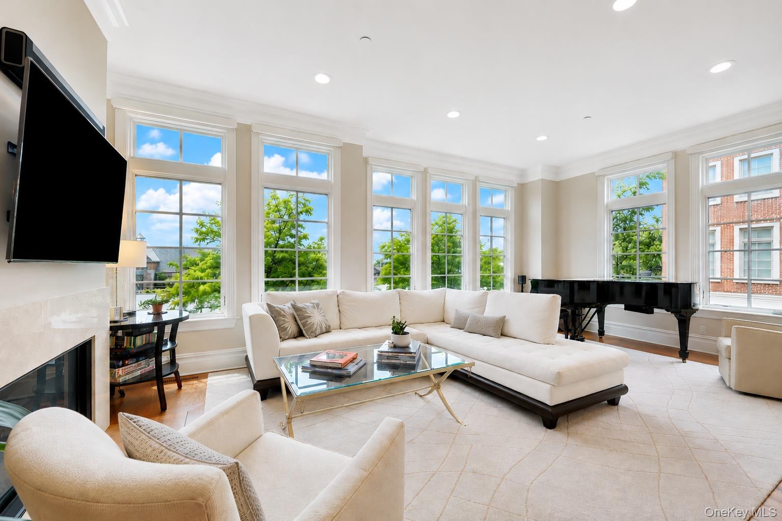 Living room with a white sectional, glass coffee table, fireplace, and grand piano beside tall windows.