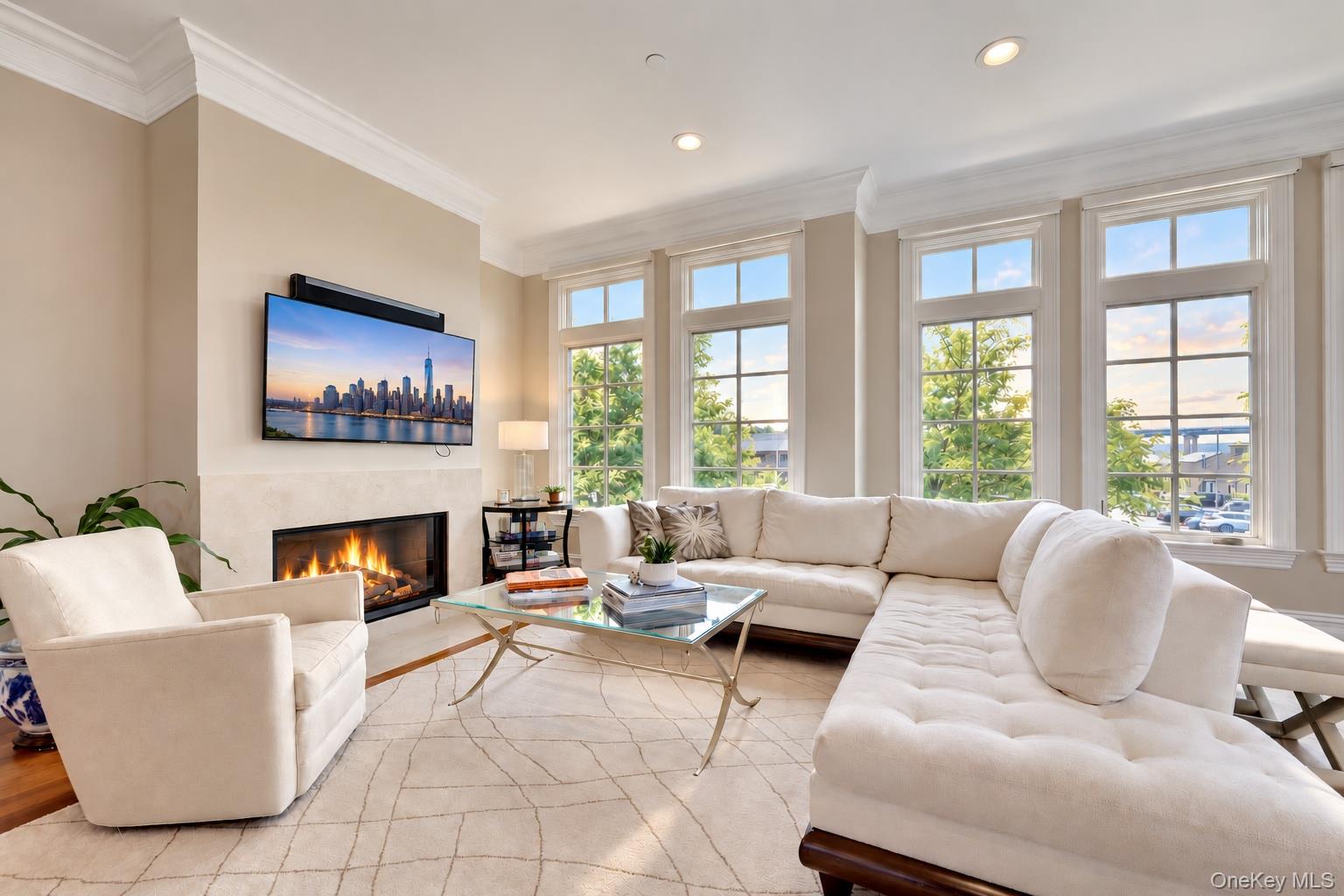 Living room with white seating, a glass coffee table, a lit fireplace, and tall windows.