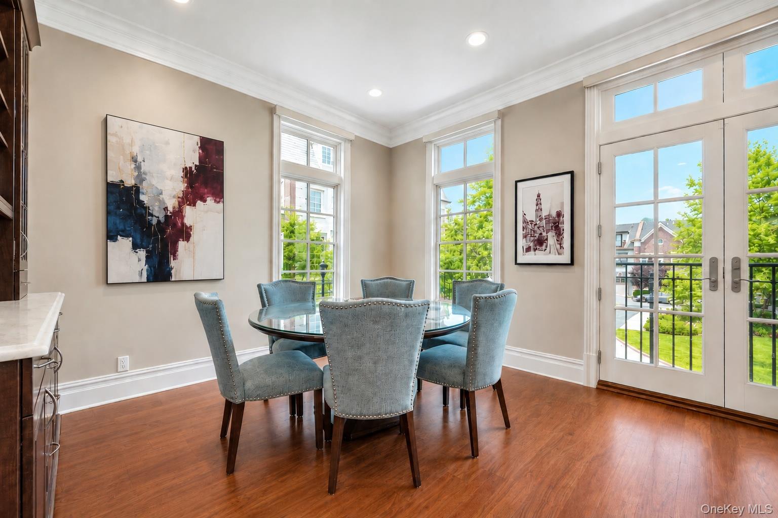 Dining room with a round glass table, blue chairs, hardwood floors, and tall windows overlooking greenery.