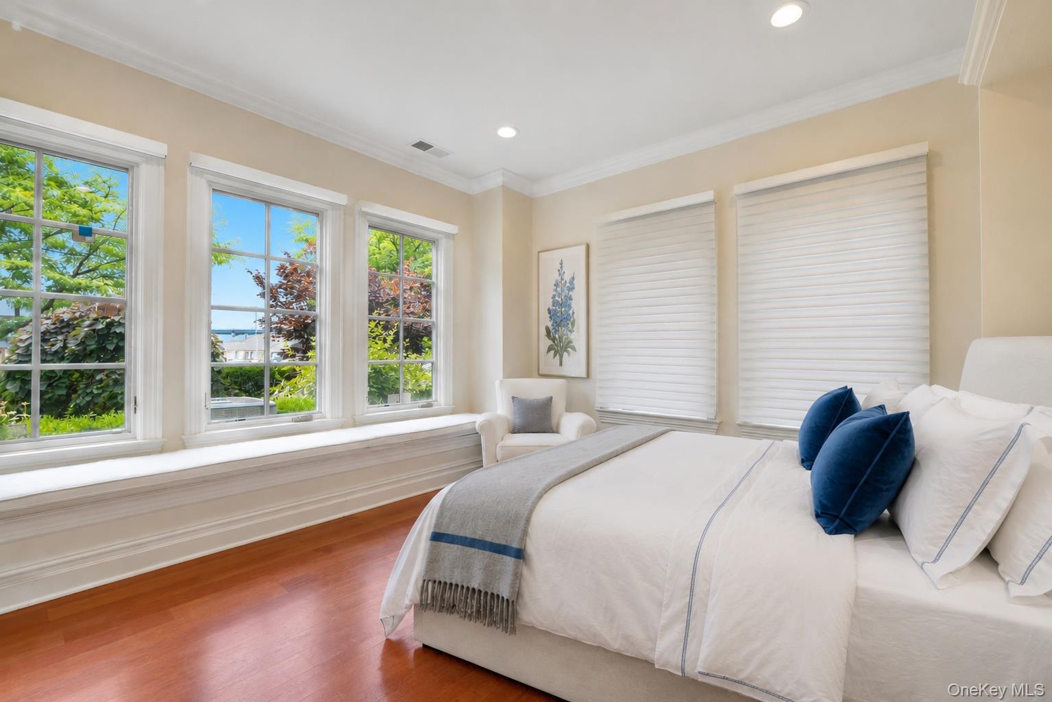 Staged bedroom with a white bed, blue pillows, and large windows overlooking greenery.