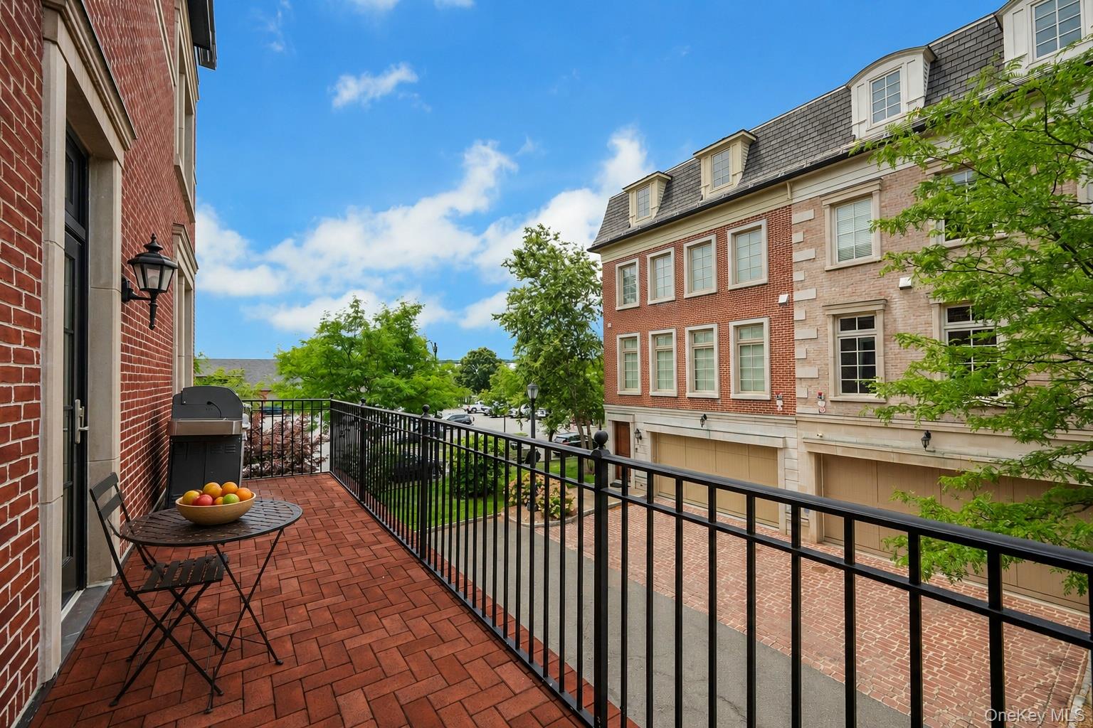 Brick balcony with black railing, outdoor table and chairs, overlooking neighboring buildings and trees.
