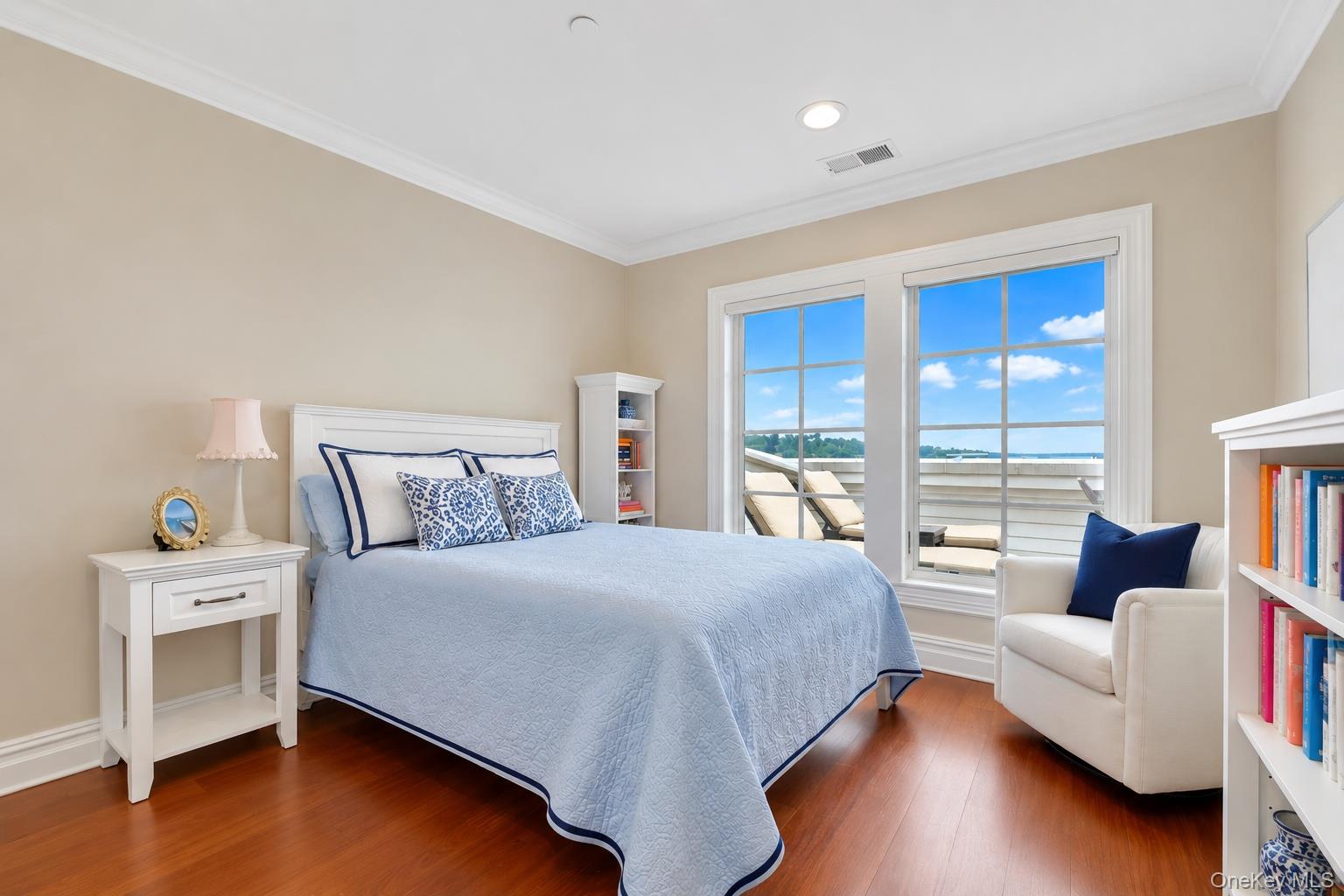 Bedroom with pale blue bedding, hardwood floors, and large windows overlooking a terrace and distant water.