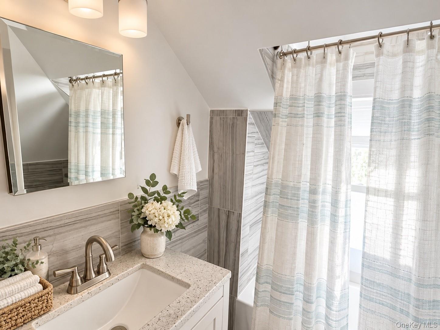 Bathroom vanity with a large mirror, gray tile, and white curtains over a bright window.