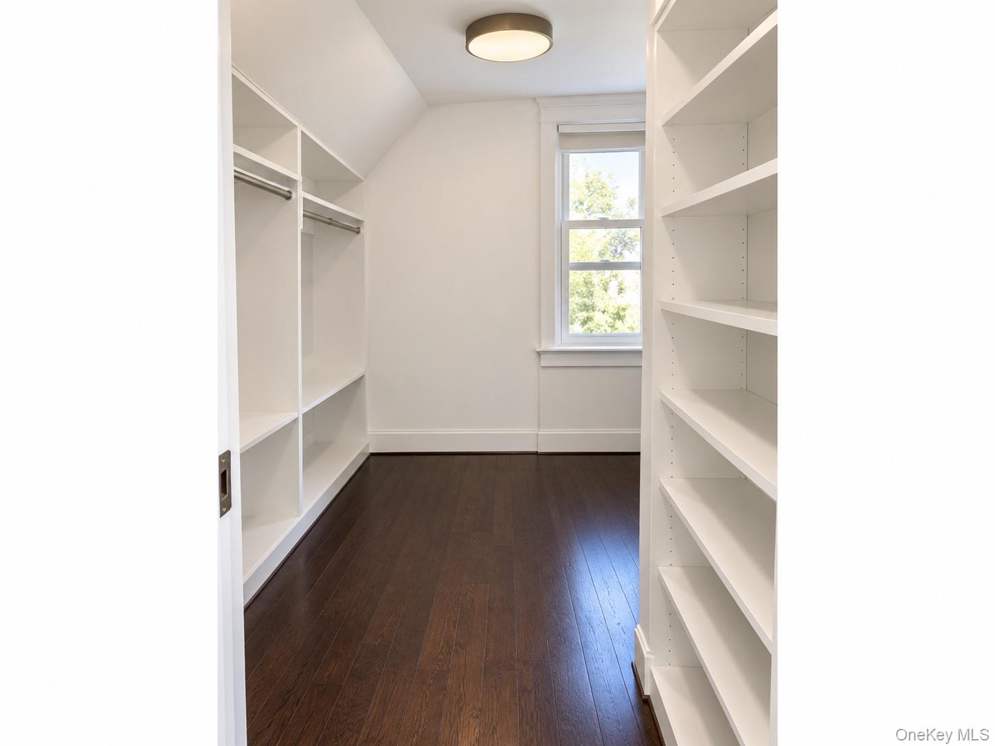 Walk-in closet with built-in shelves, hanging rods, dark wood flooring, and a window.