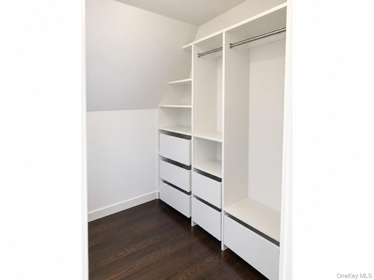 Walk-in closet with white built-in drawers and shelving, metal hanging rods, and dark hardwood flooring.