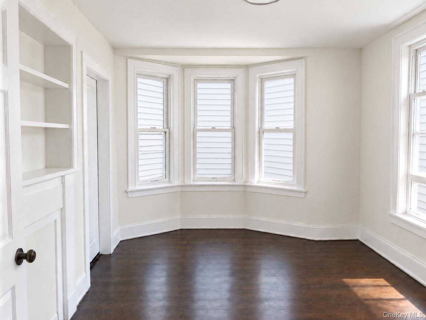 Empty room with dark hardwood floors, built-in shelves, and three bay windows.