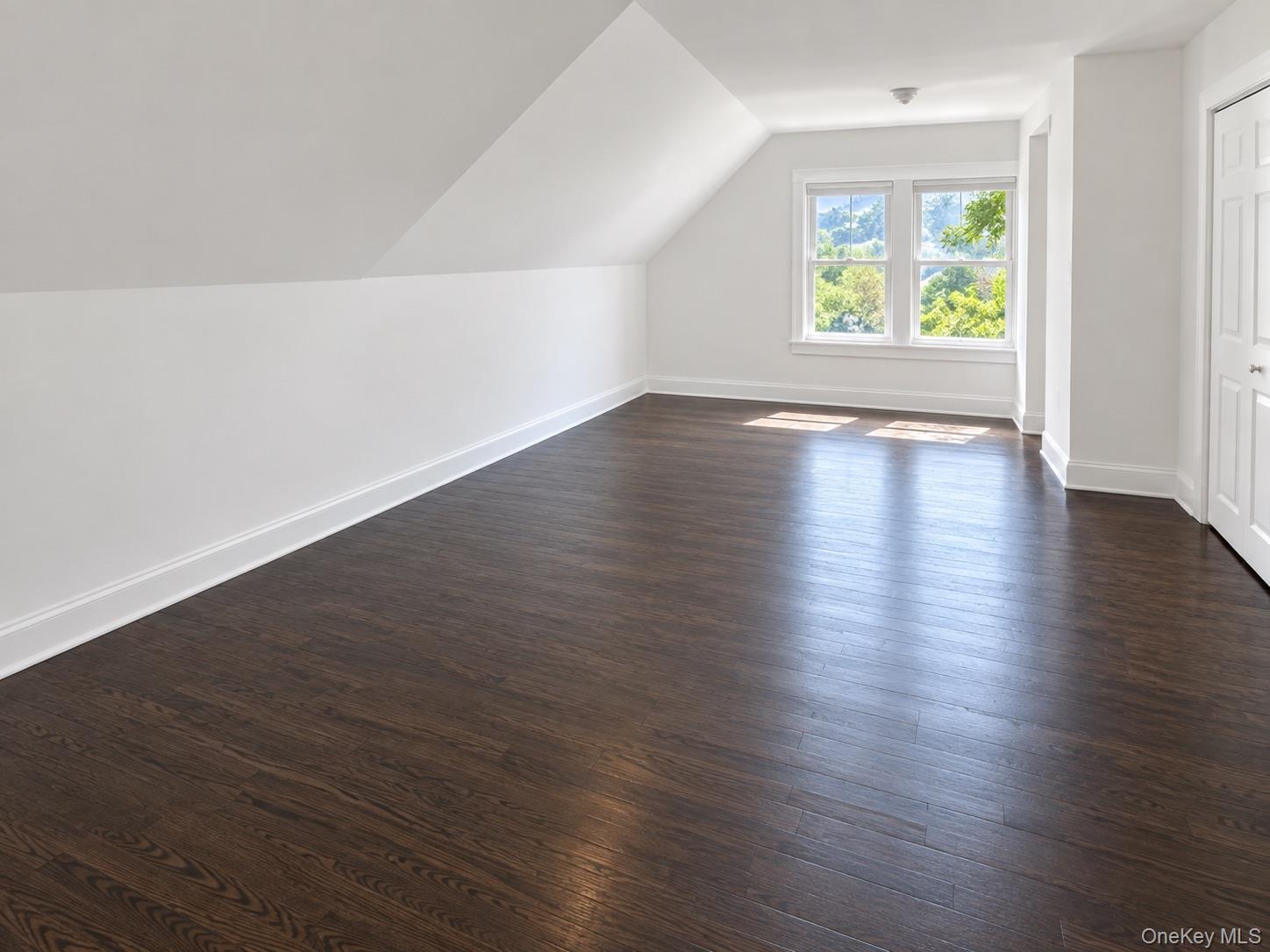 Empty finished attic room with dark wood floors, sloped ceilings, and a sunny window overlooking trees.