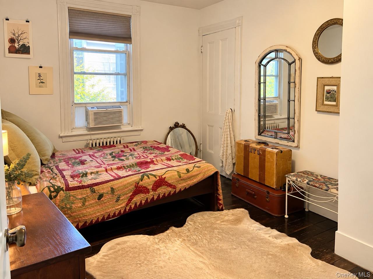 Bedroom with a floral quilt, dark hardwood floors, and vintage trunks beneath an arched mirror.