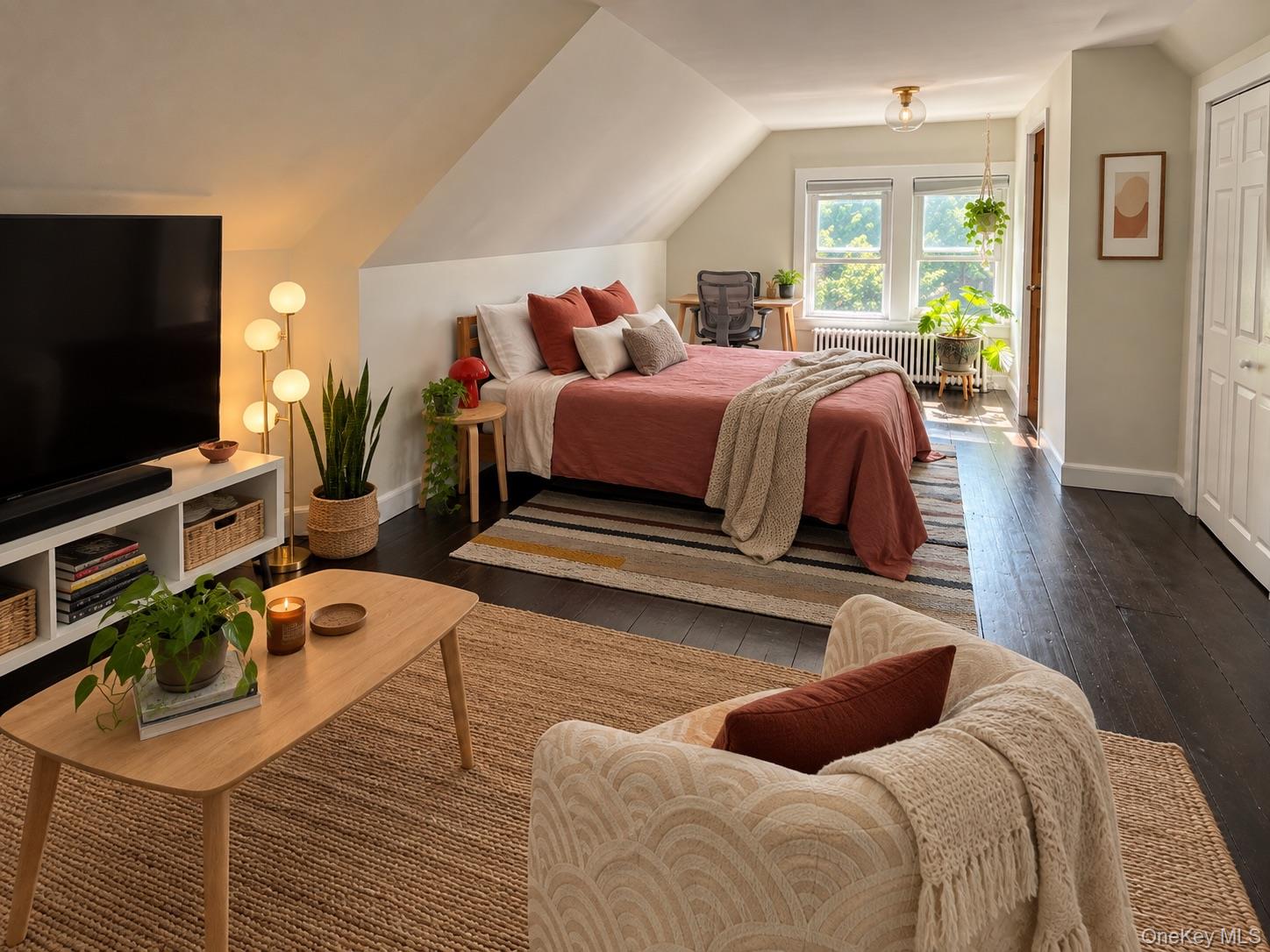 Staged attic bedroom with terracotta bedding, sloped ceilings, dark floors, and a desk by two windows.