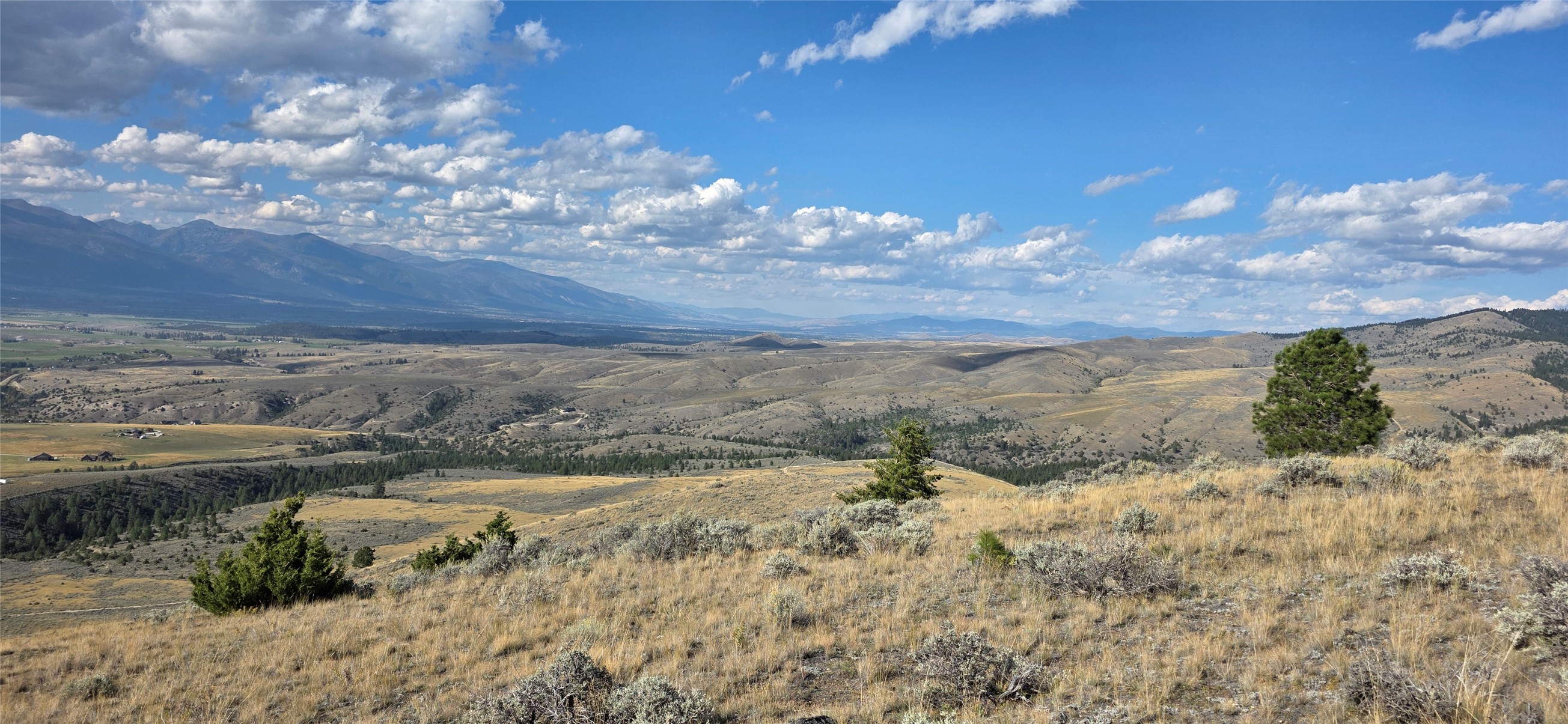 Dry hillside lot overlooking rolling valleys and distant mountains beneath a cloud-filled blue sky.
