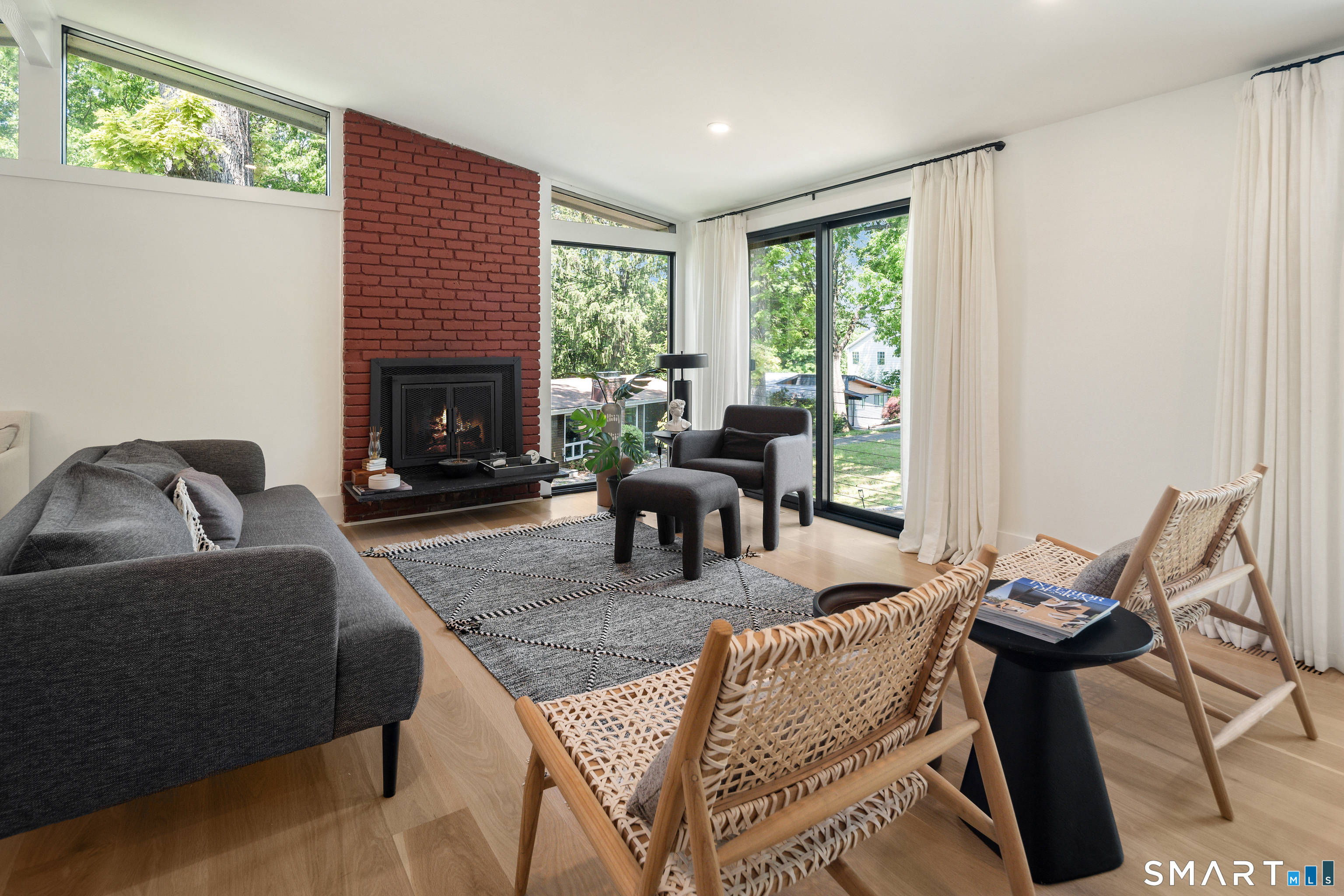 Furnished living room with a brick fireplace, woven chairs, and large windows overlooking trees.