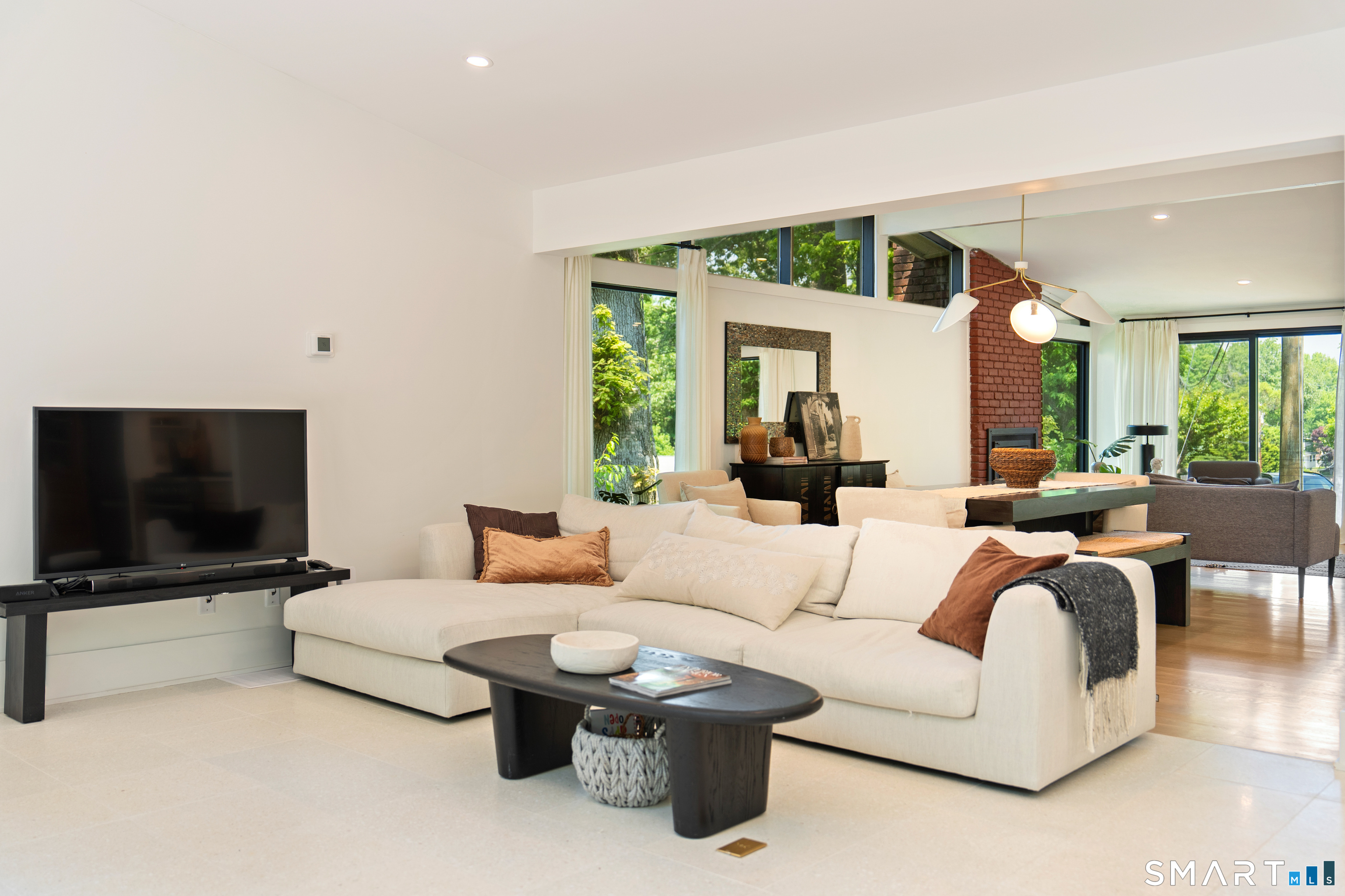Furnished living room with a cream sectional, tall windows overlooking trees, and a red brick fireplace.