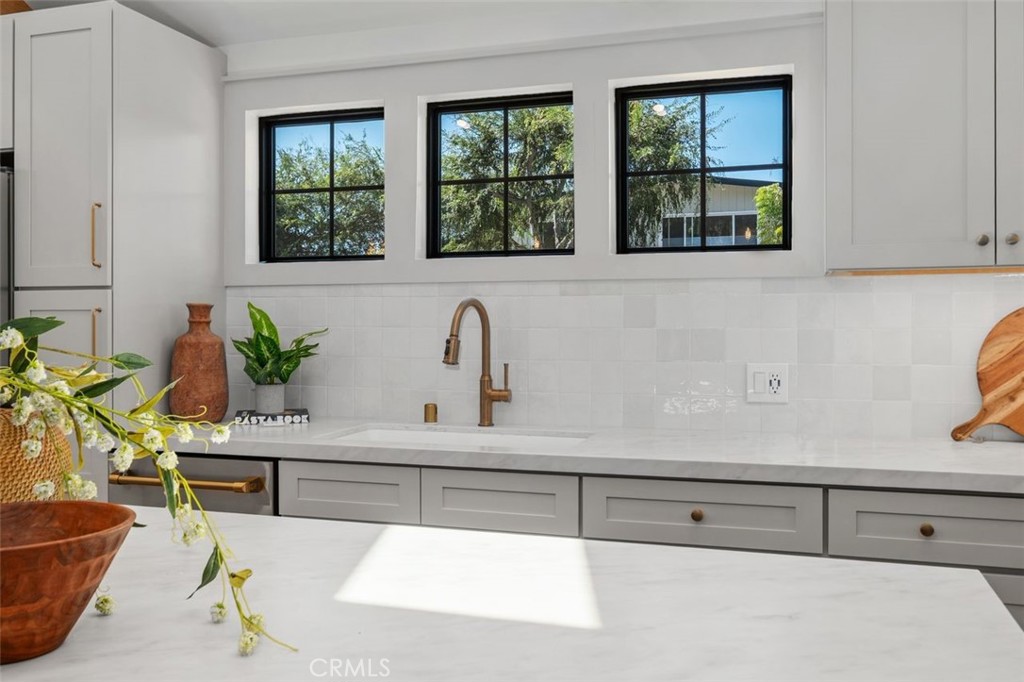 Bright kitchen with gray cabinetry, marble counters, brass faucet, and three windows overlooking trees.