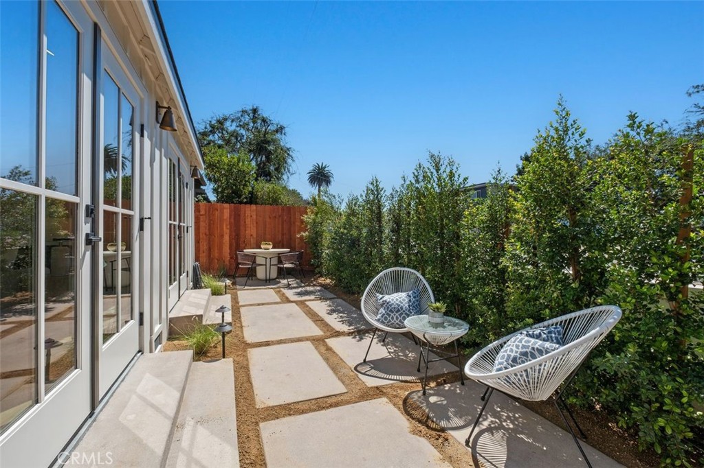 Furnished patio with pale stone pavers, white-framed glass doors, and wire lounge chairs beside green hedges.