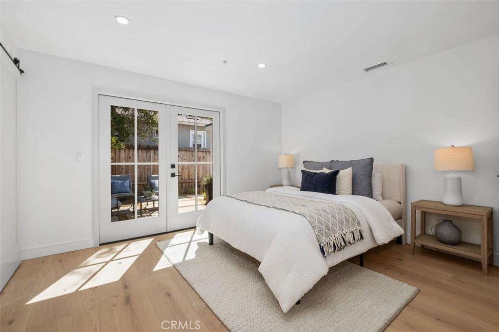 Staged bedroom with white bedding, light wood floors, and French doors opening to a patio.