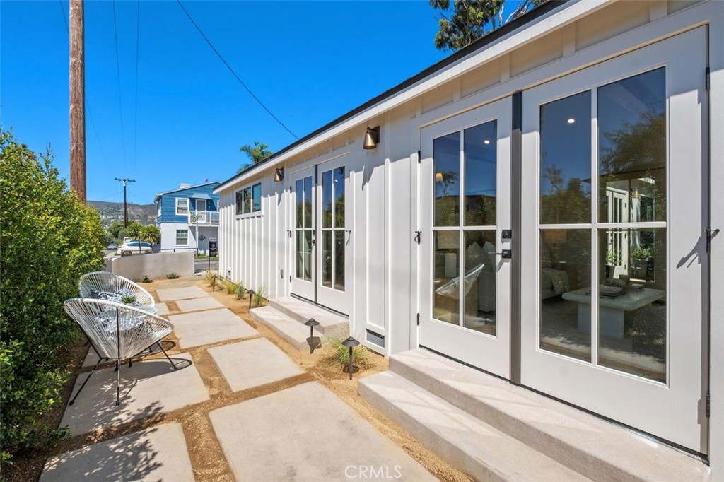 Modern white home with tall glass doors beside a paved patio and metal chairs.