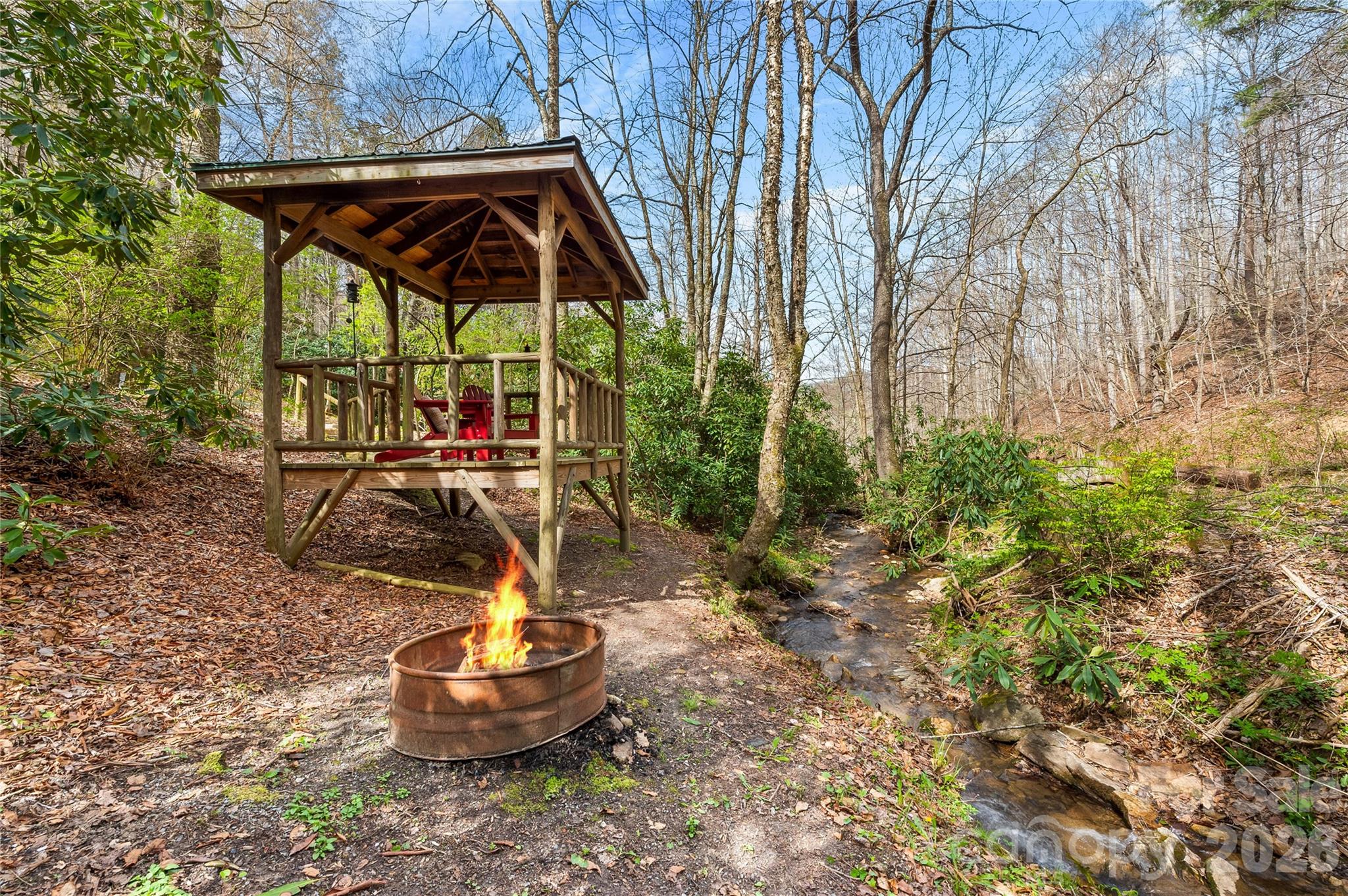 Wooden gazebo and fire pit beside a rocky creek in a wooded setting.