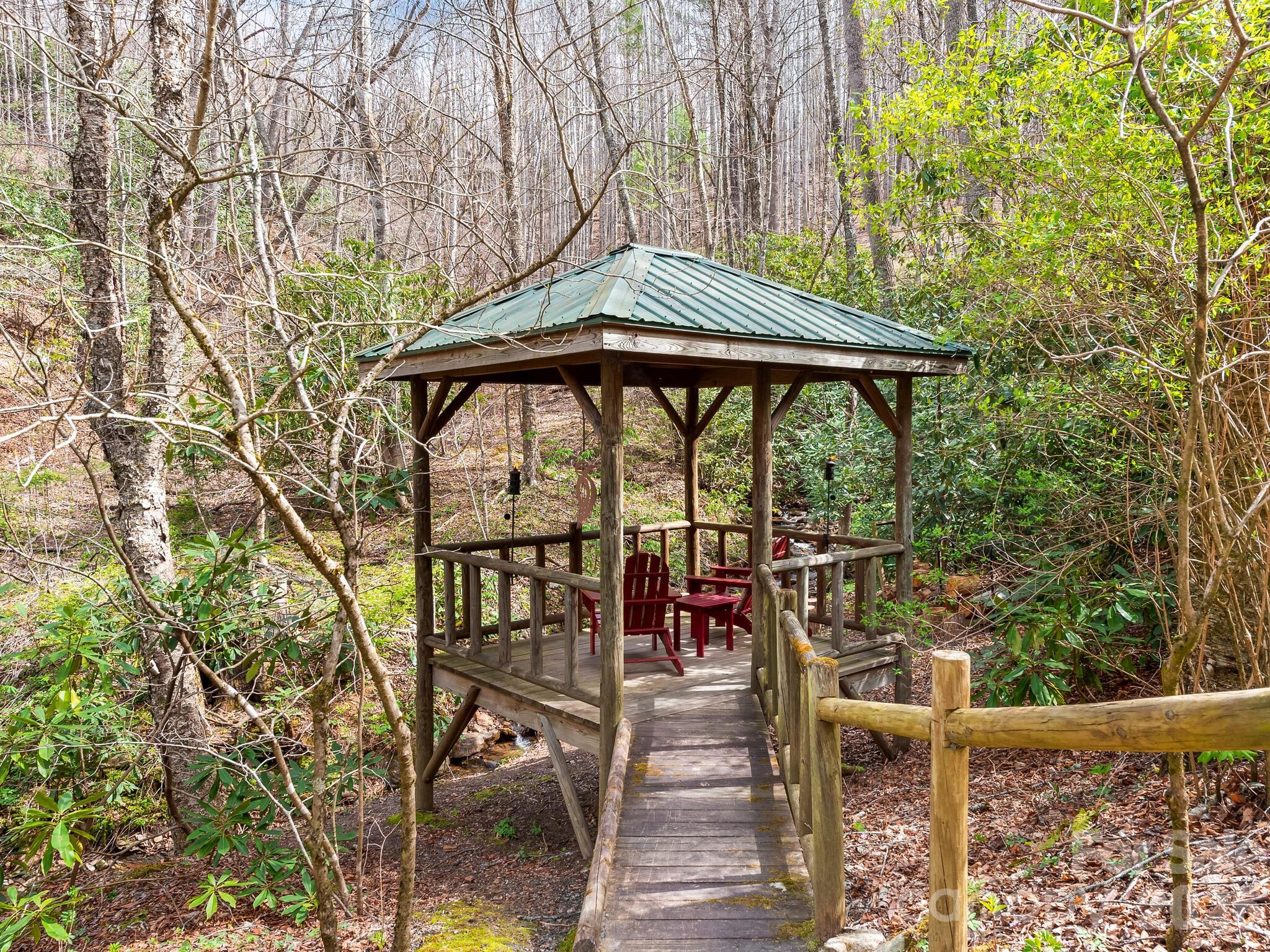 Woodland gazebo with red chairs, a green roof, and a wooden walkway beside a creek.
