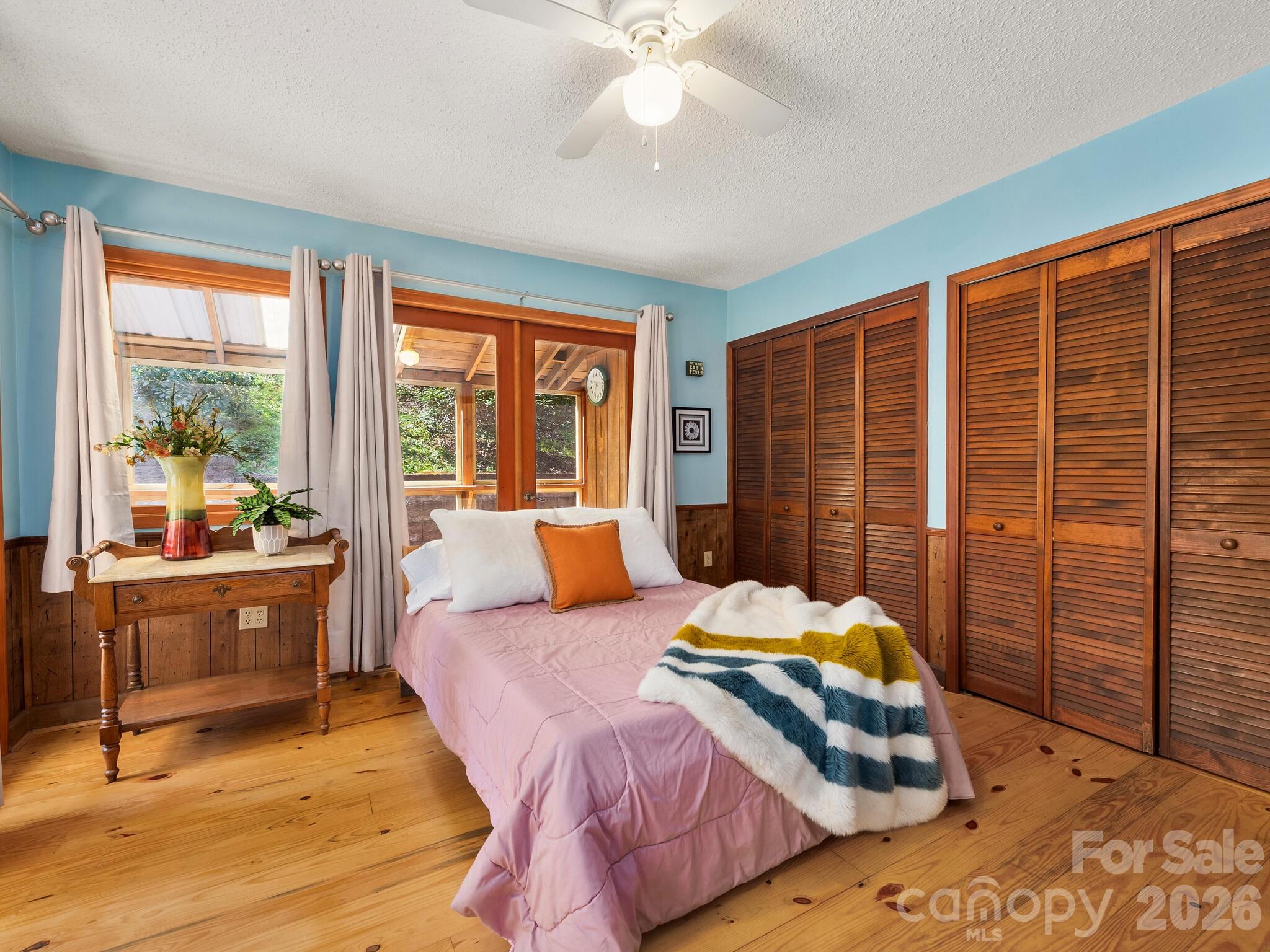 Bedroom with a pink-covered bed, striped throw, wooden floors, and louvered closet doors.