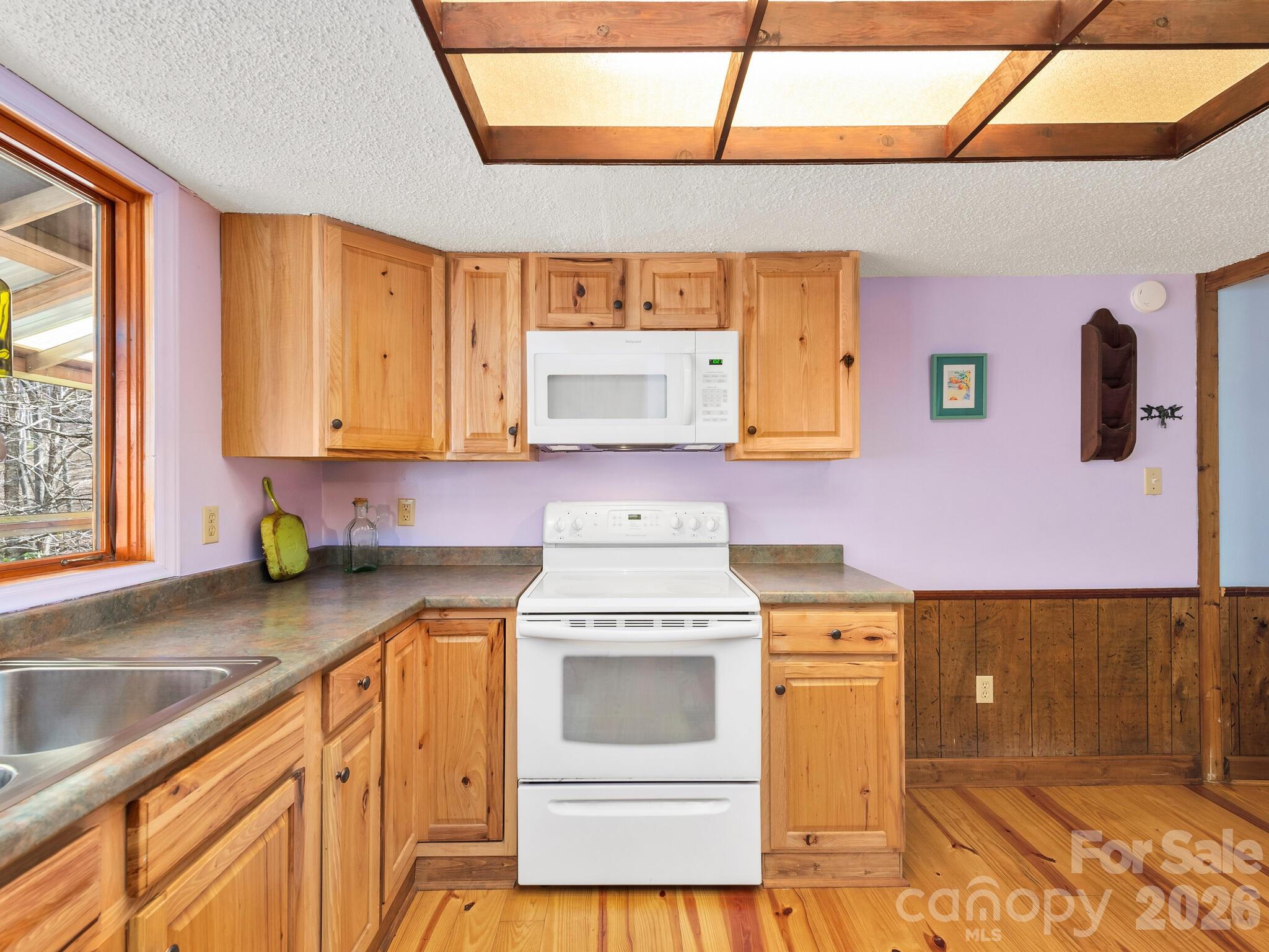 Kitchen with knotty wood cabinets and floors, white range and microwave, and a large window.