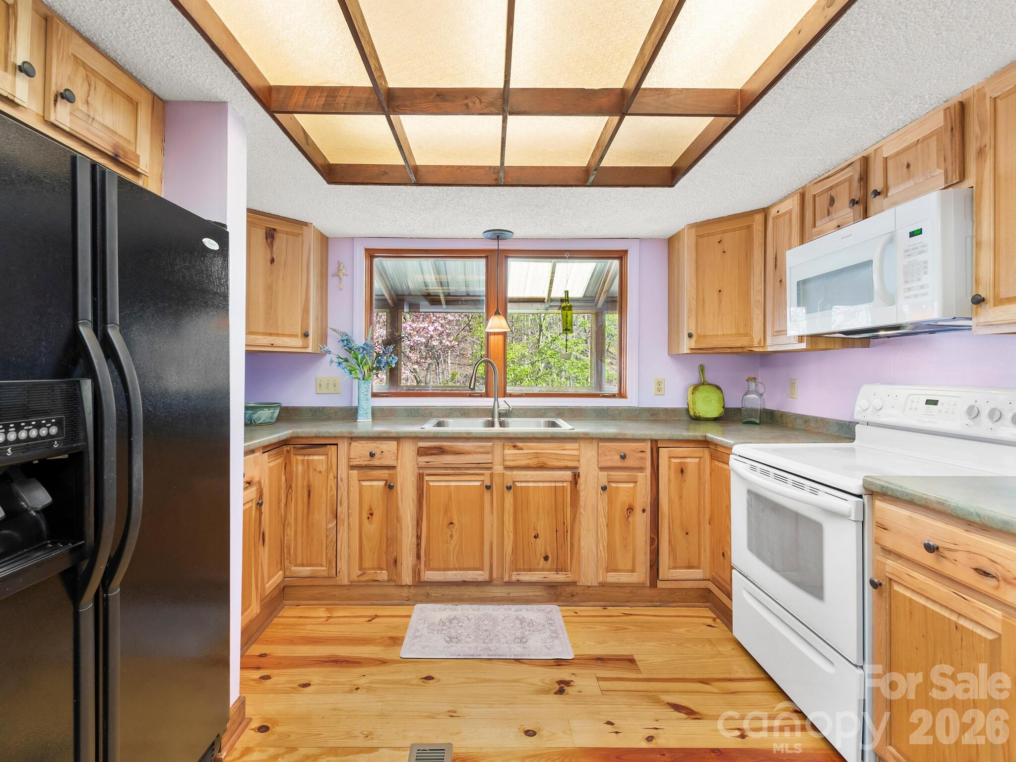 Kitchen with natural wood cabinets, plank floors, and a sink beneath a window overlooking trees.