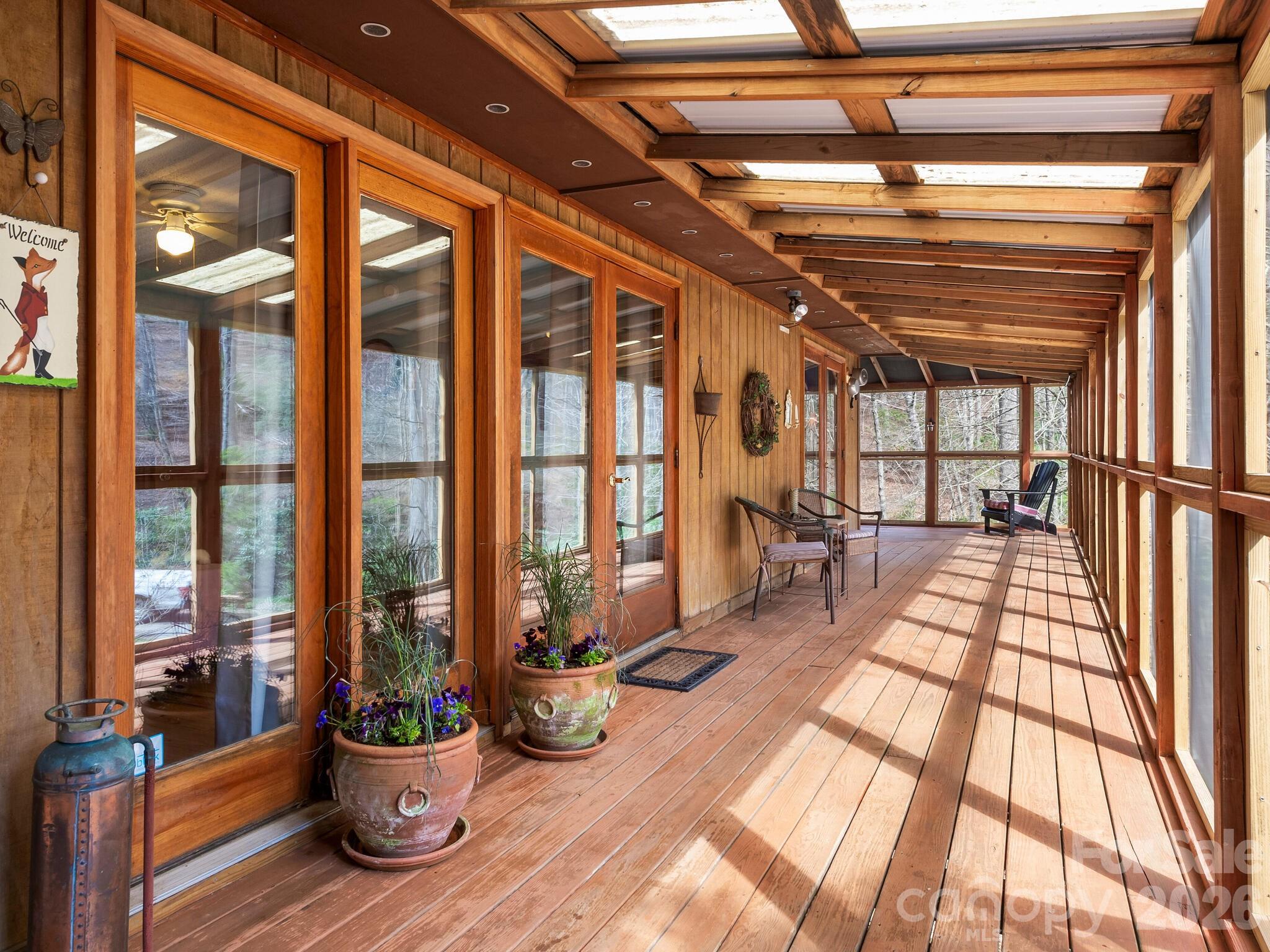 Covered wooden porch with skylight panels, potted flowers, and seating beside large windows.