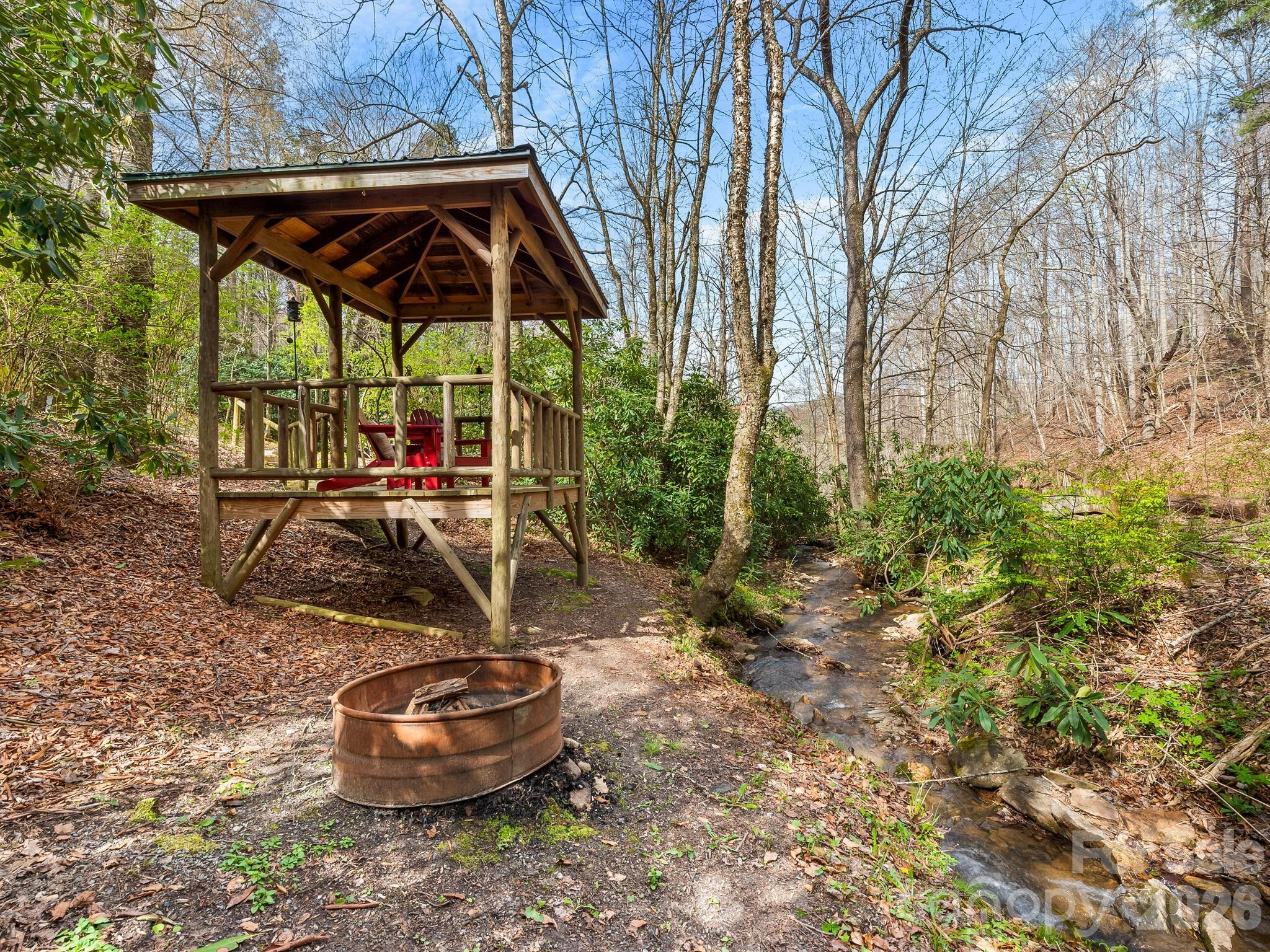 Wooden gazebo with red chairs beside a creek, with a metal fire pit in the foreground.