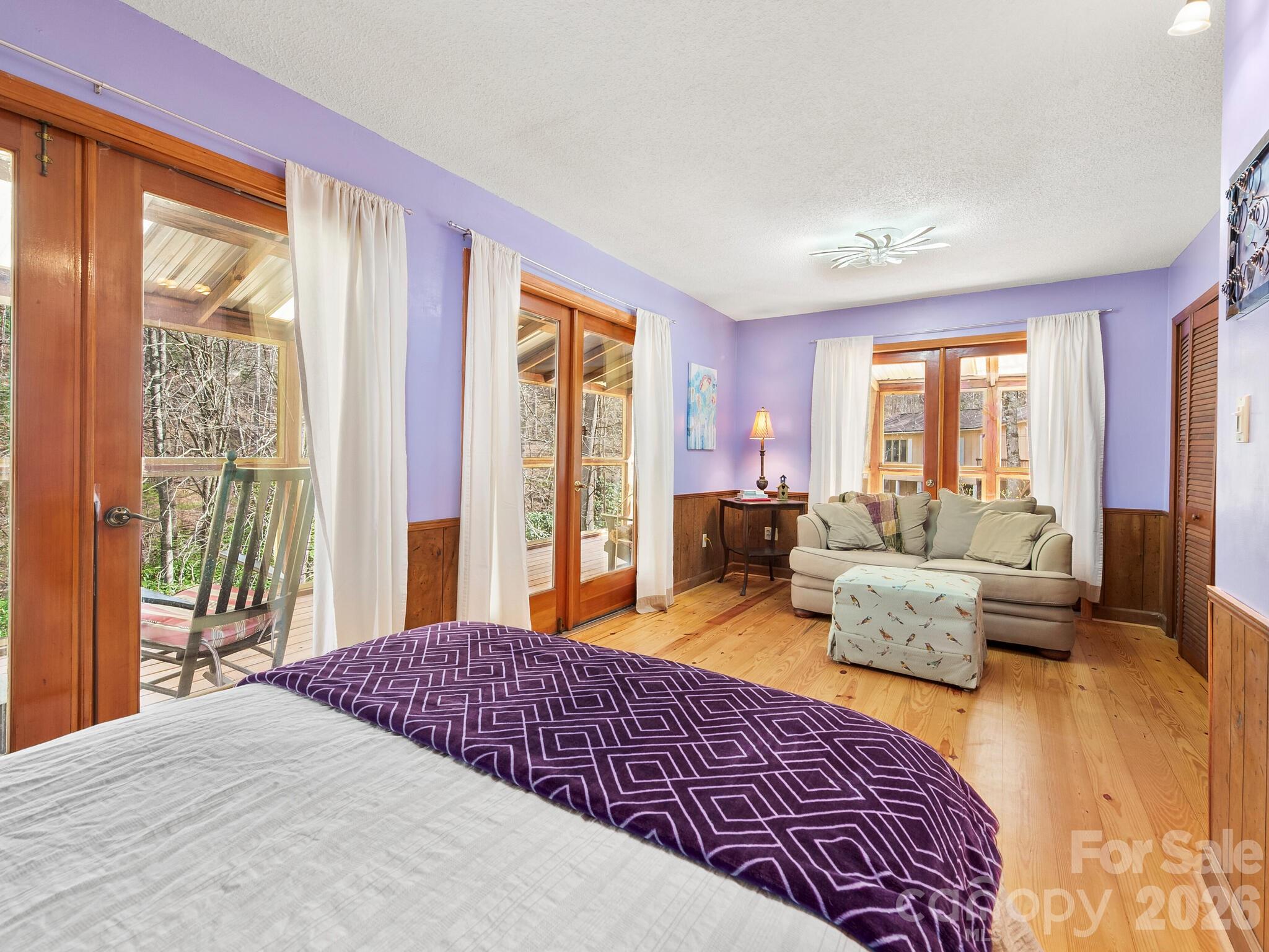 Bedroom with a sofa sitting area, purple walls, wood floors, and glass doors opening to a wooded deck.