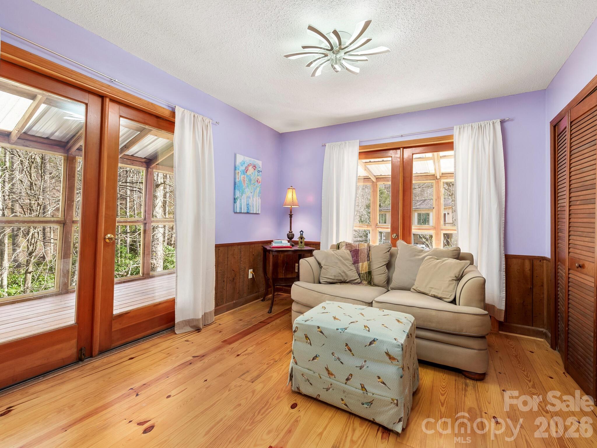 Living room with lavender walls, wood floors, and glass doors overlooking trees.