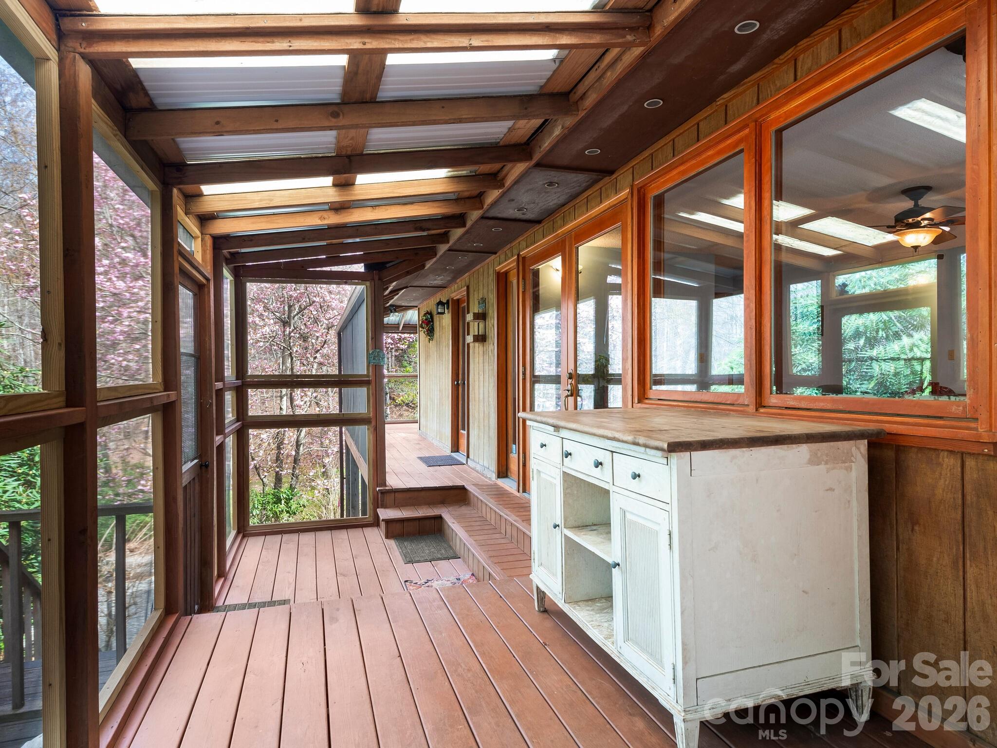 Covered wooden porch with exposed beams, large windows, and a white cabinet with a countertop.
