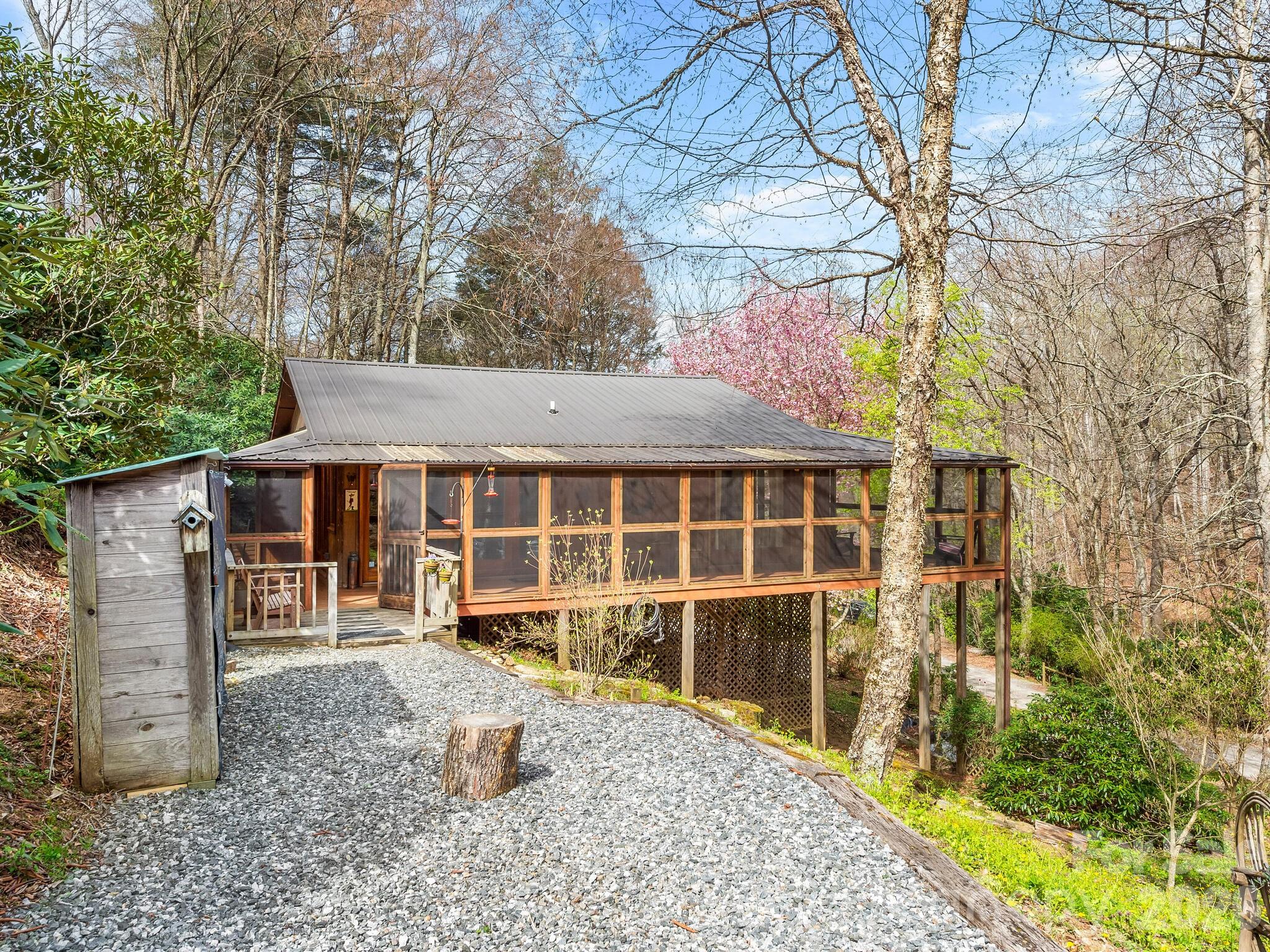 Raised cabin with a long screened porch, metal roof, and gravel driveway among wooded spring surroundings.