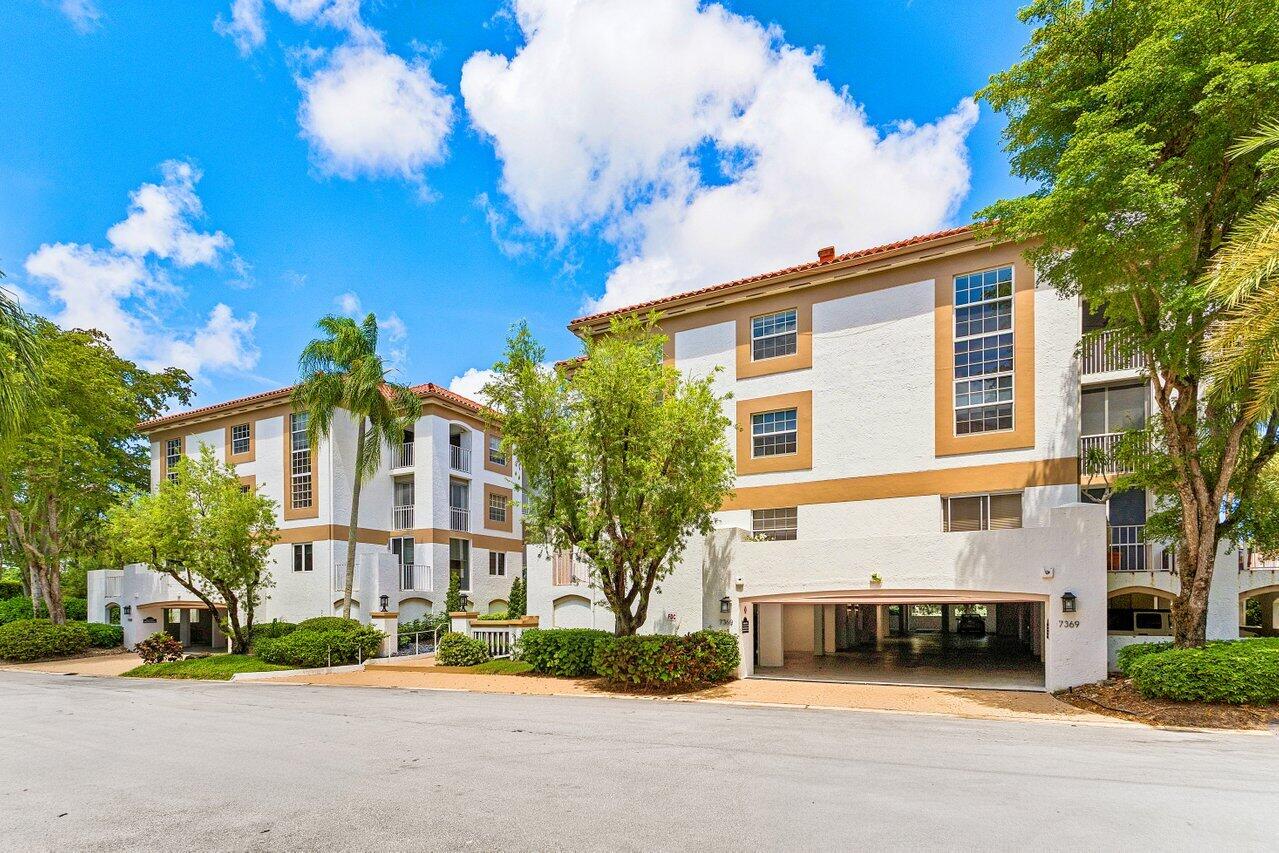Multi-story condominium building with tan trim, balconies, covered parking, and tropical landscaping.