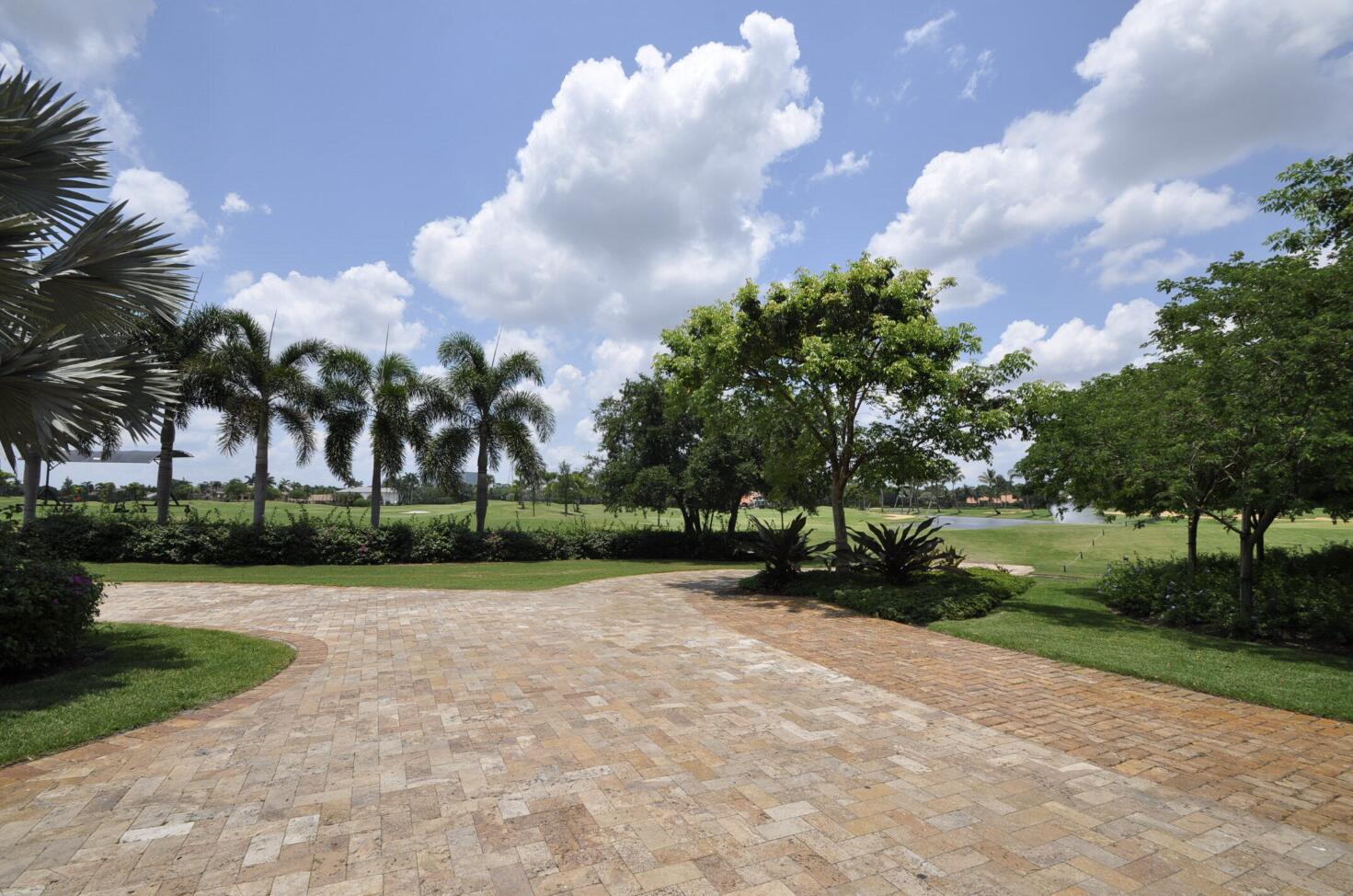 Paver driveway bordered by palms and lawn, with a golf course and pond beyond.
