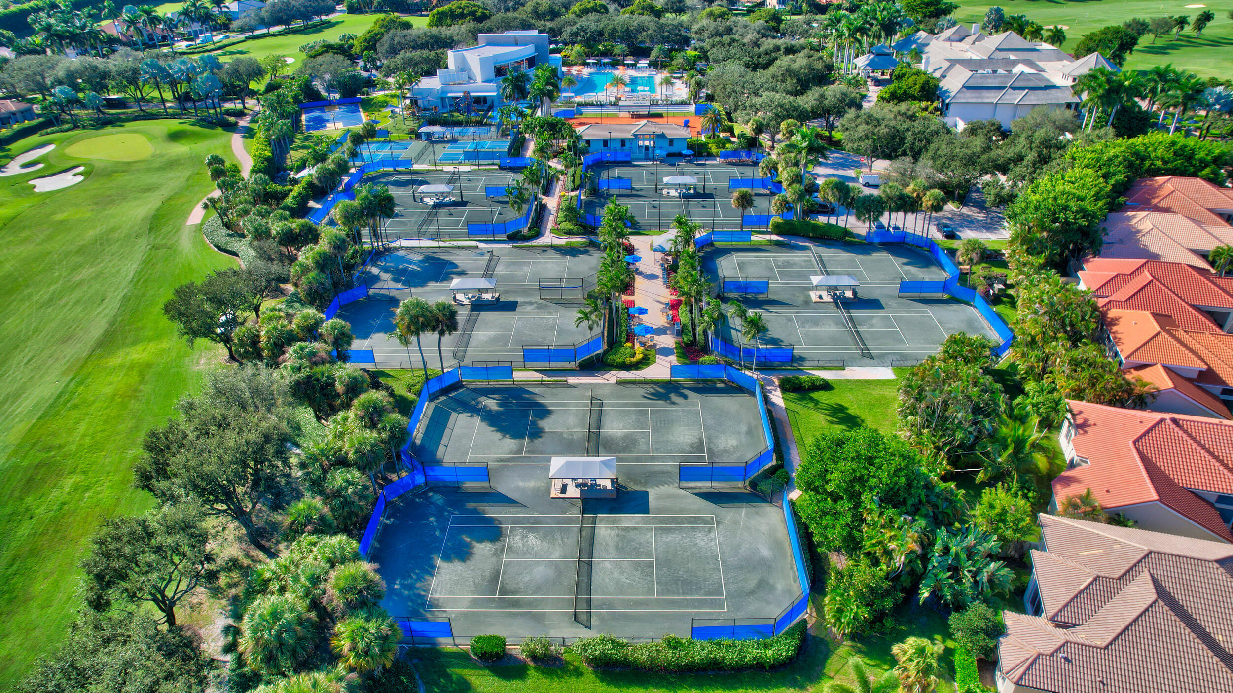Aerial view of multiple tennis courts beside a golf course, pool, clubhouse, and palm-lined homes.