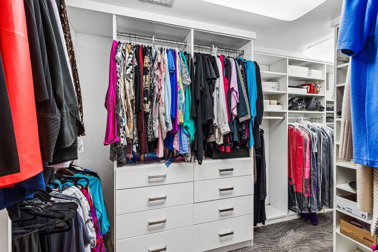 Walk-in closet with white built-ins, crowded clothing racks, shoe shelves, and gray carpet.