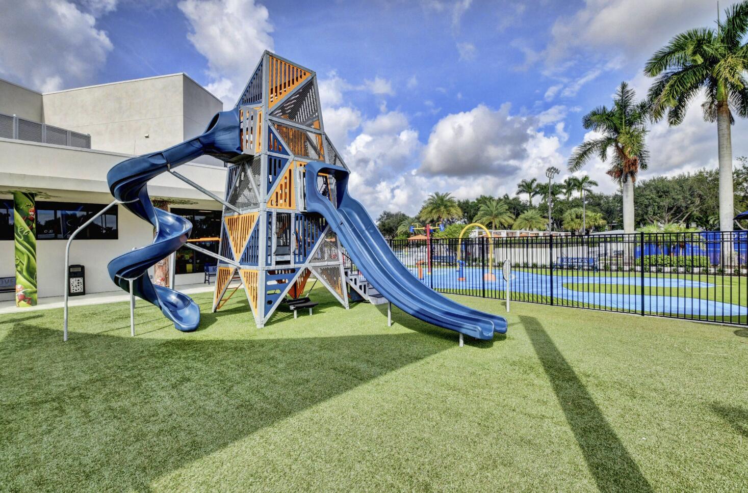 Community playground with blue slides and a fenced splash pad beside a clubhouse.