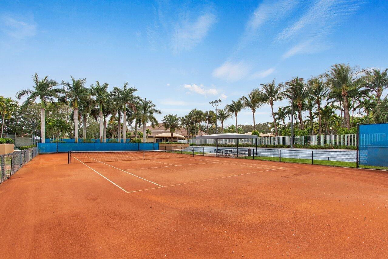 Red clay tennis court with a net, palm trees, and a second court beyond the fence.