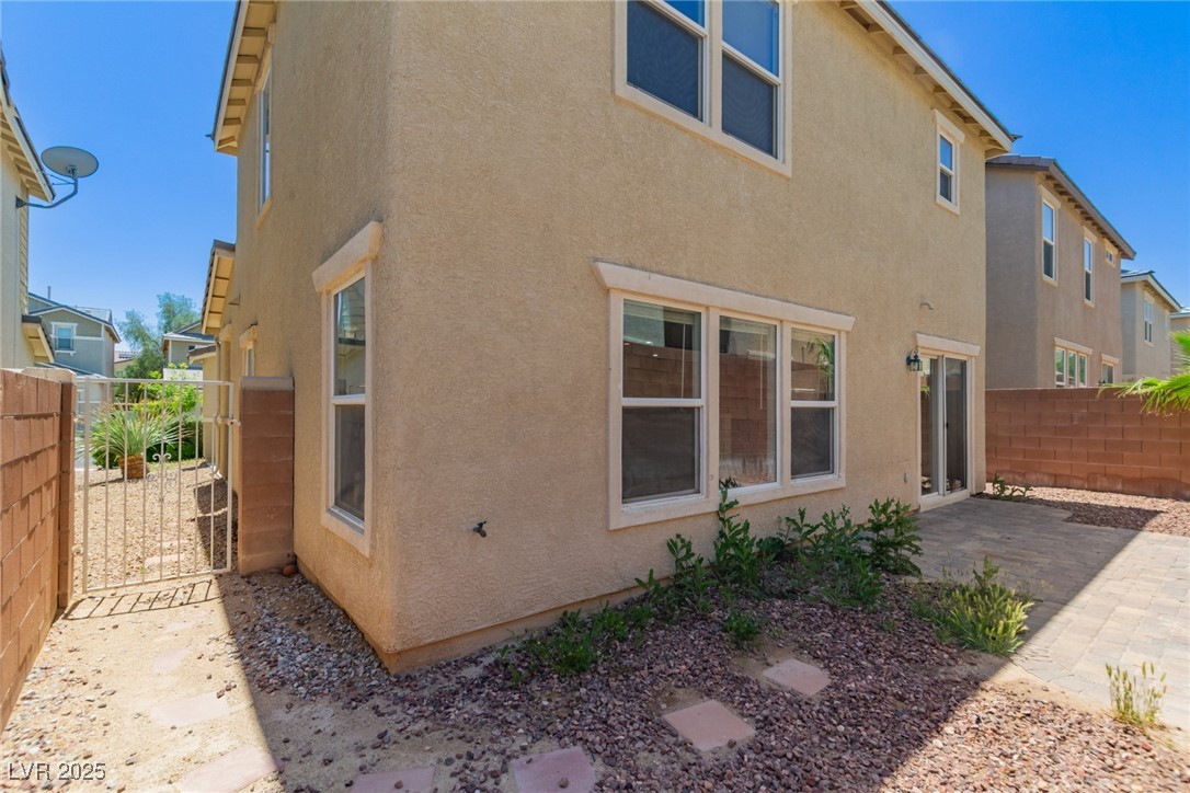 Two-story tan stucco house with a side gate, rock landscaping, and a paved patio.