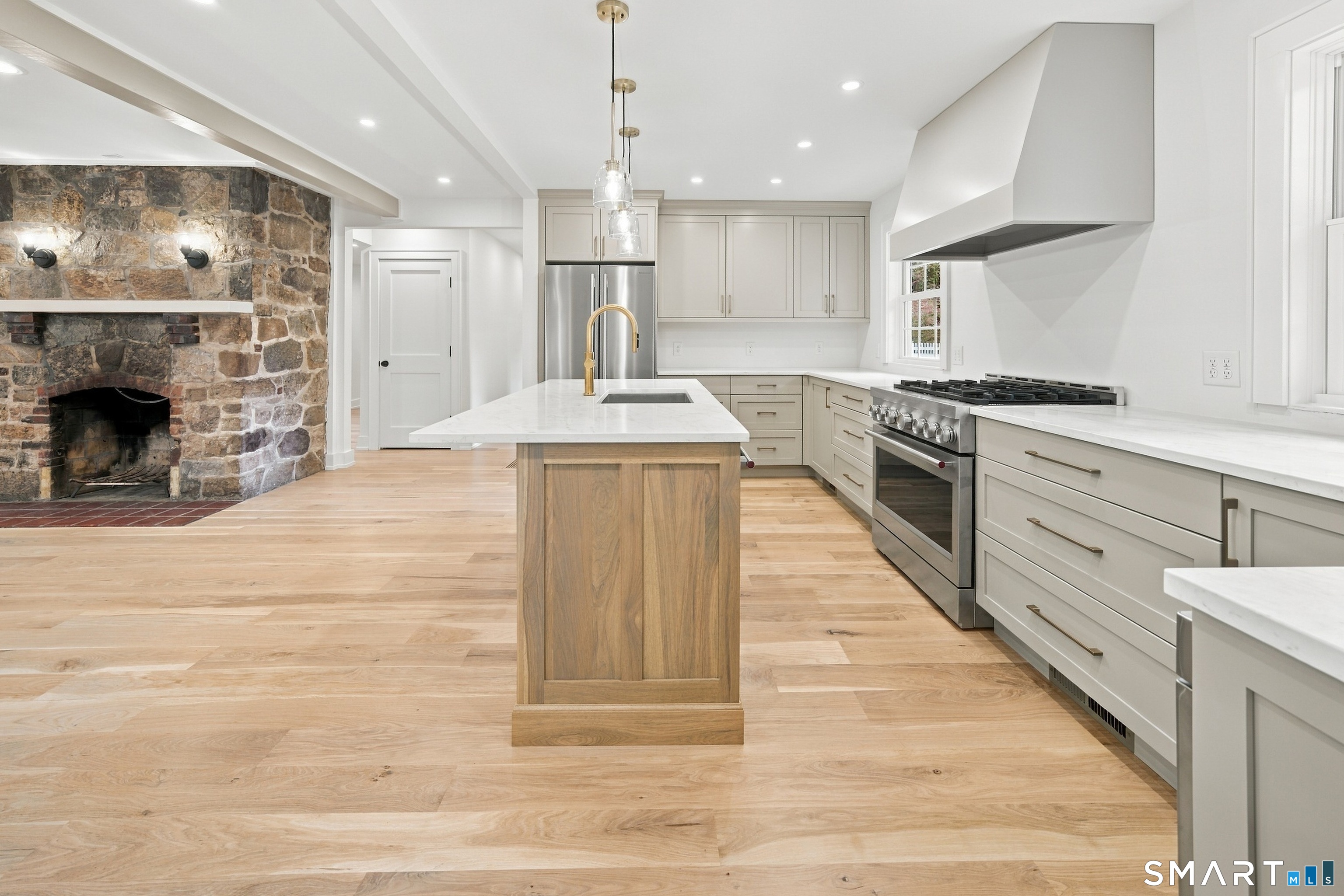 Renovated kitchen with a wood island, stainless steel range, pale wood floors, and a large stone fireplace.