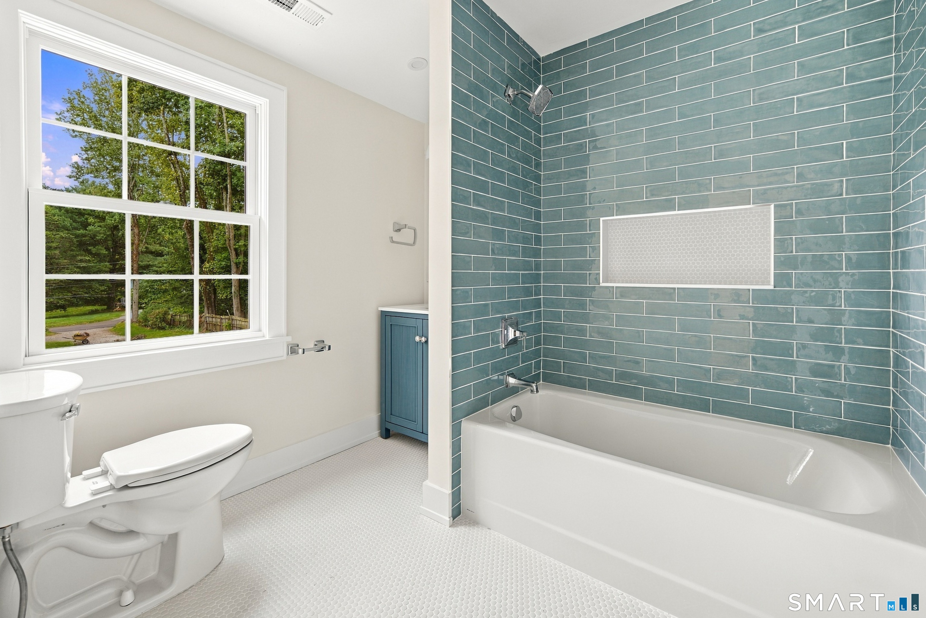 Bathroom with a teal subway tile tub surround, large window, and white penny tile floor.