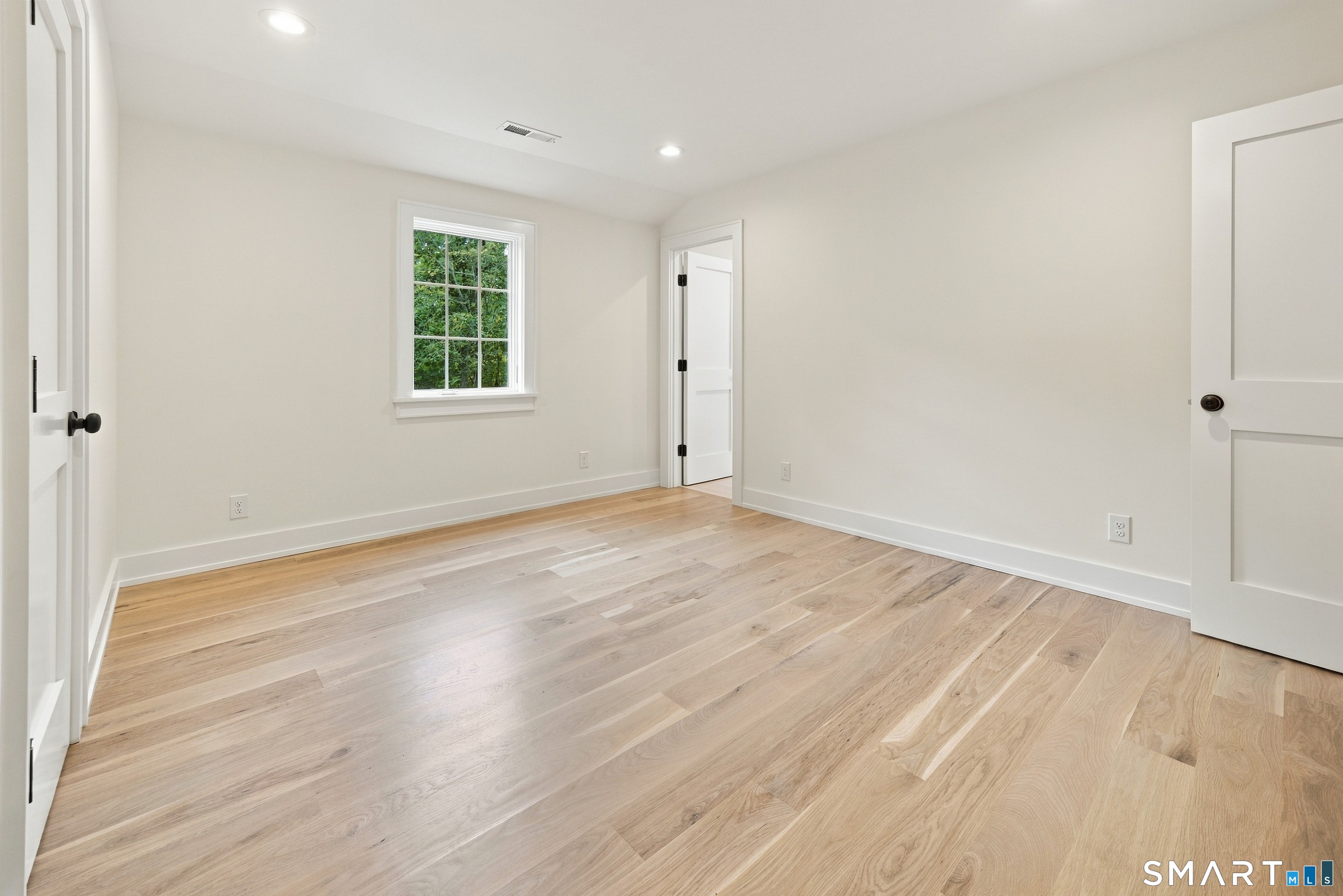 Empty room with light hardwood floors, white walls, a divided-light window, and open paneled doors.