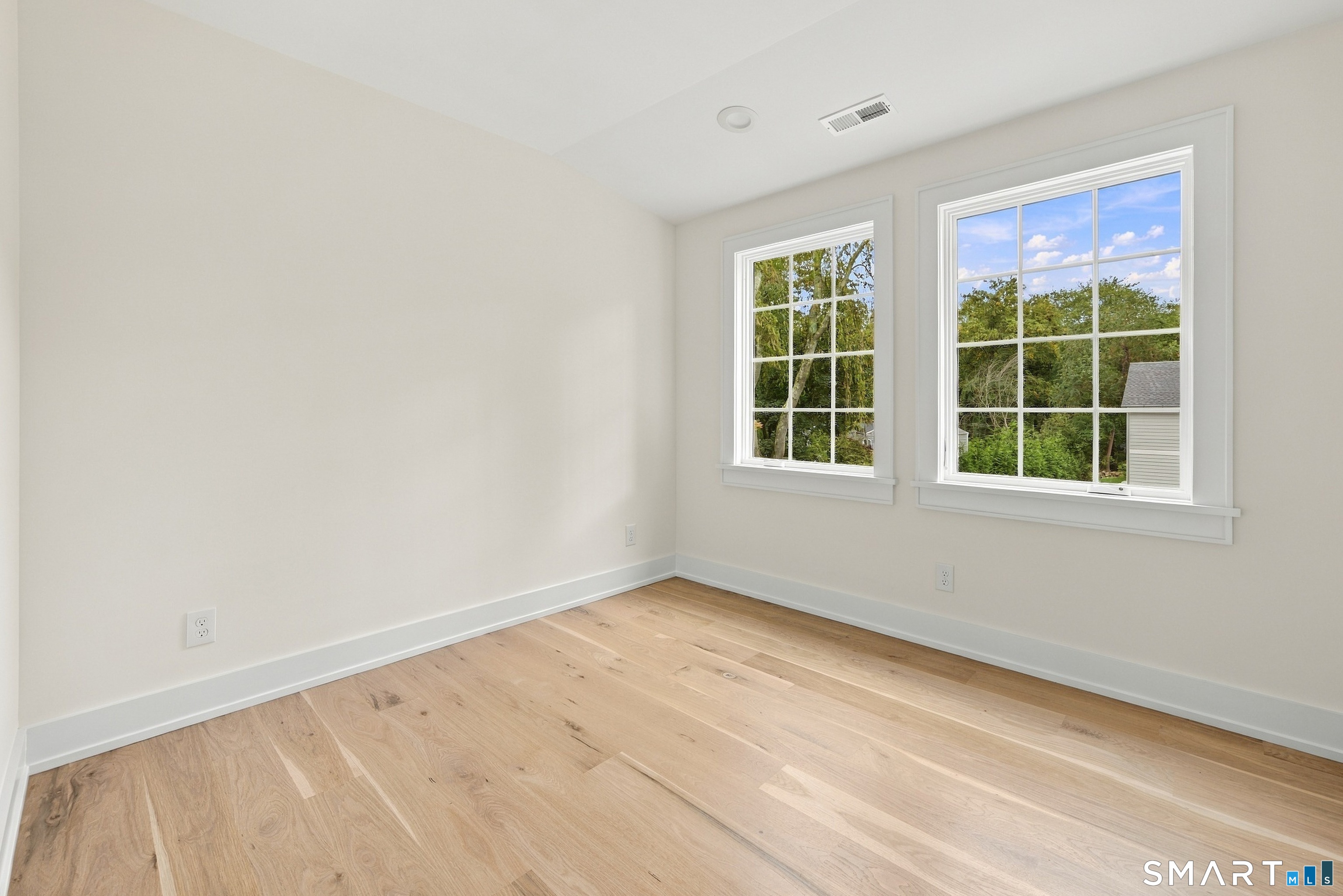 Vacant room with light wood floors and two large windows overlooking trees.