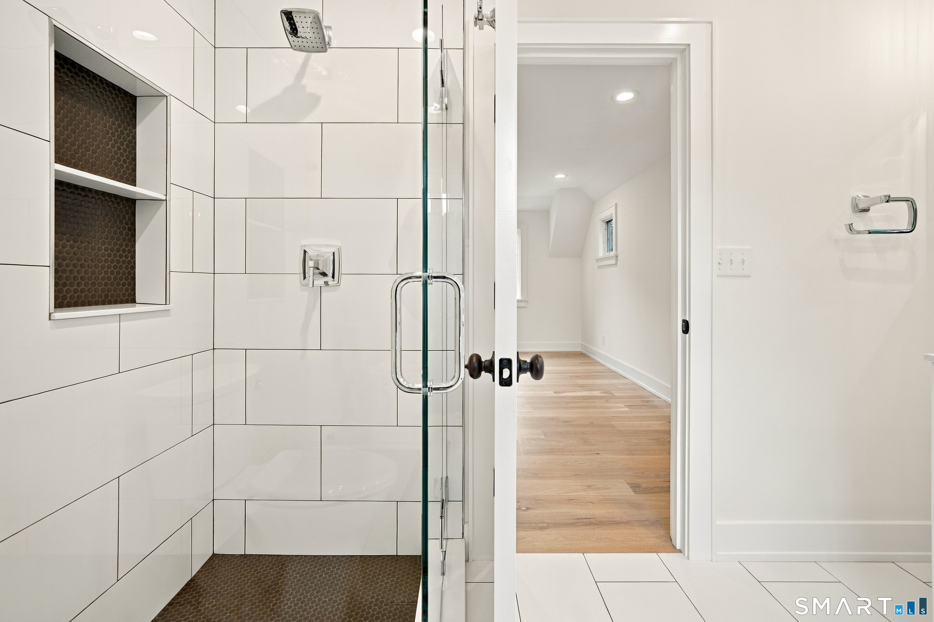 Modern tiled bathroom with a glass shower and dark mosaic niches, opening to a wood-floored hallway.