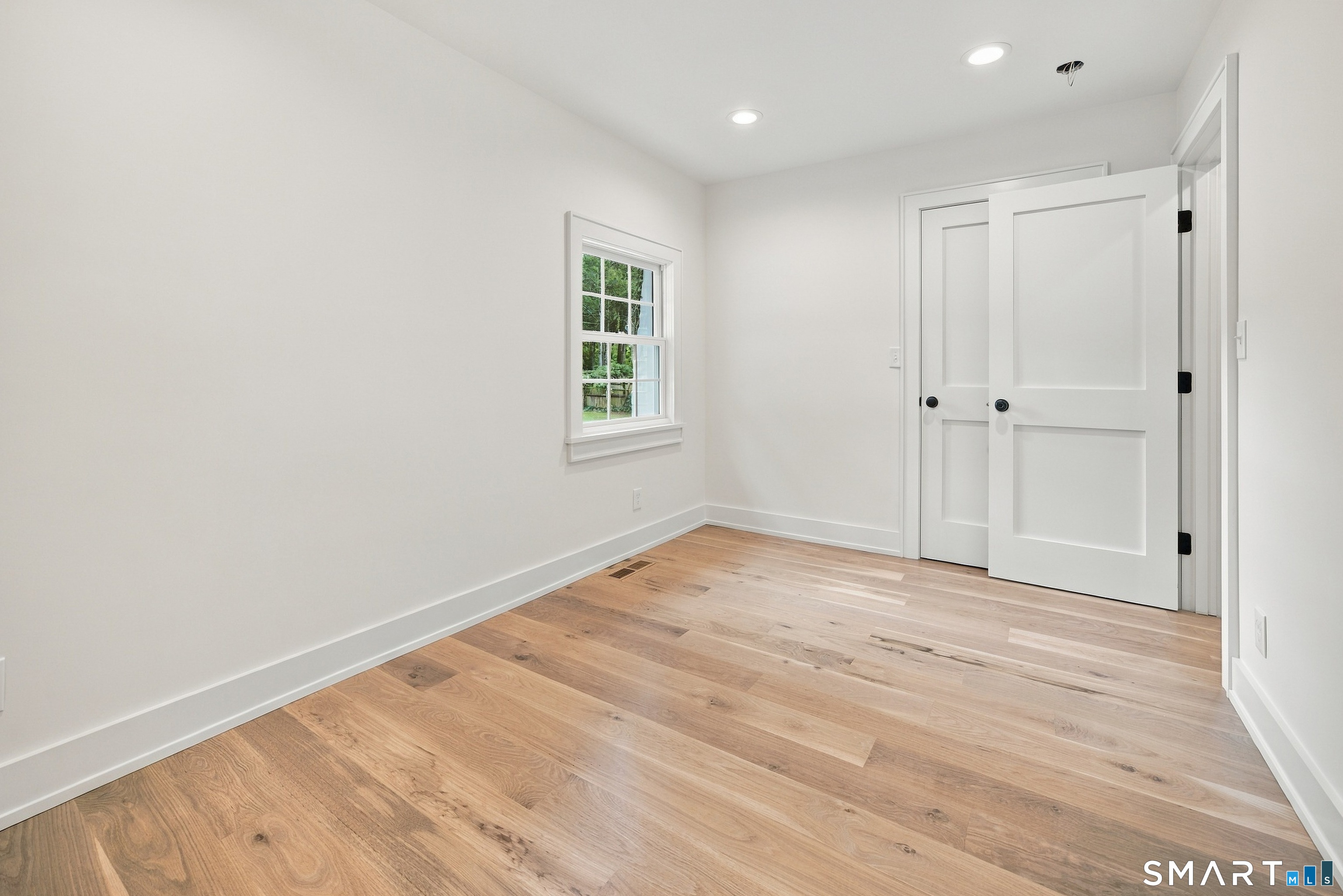 Empty room with light oak flooring, a divided-pane window, and white paneled doors.