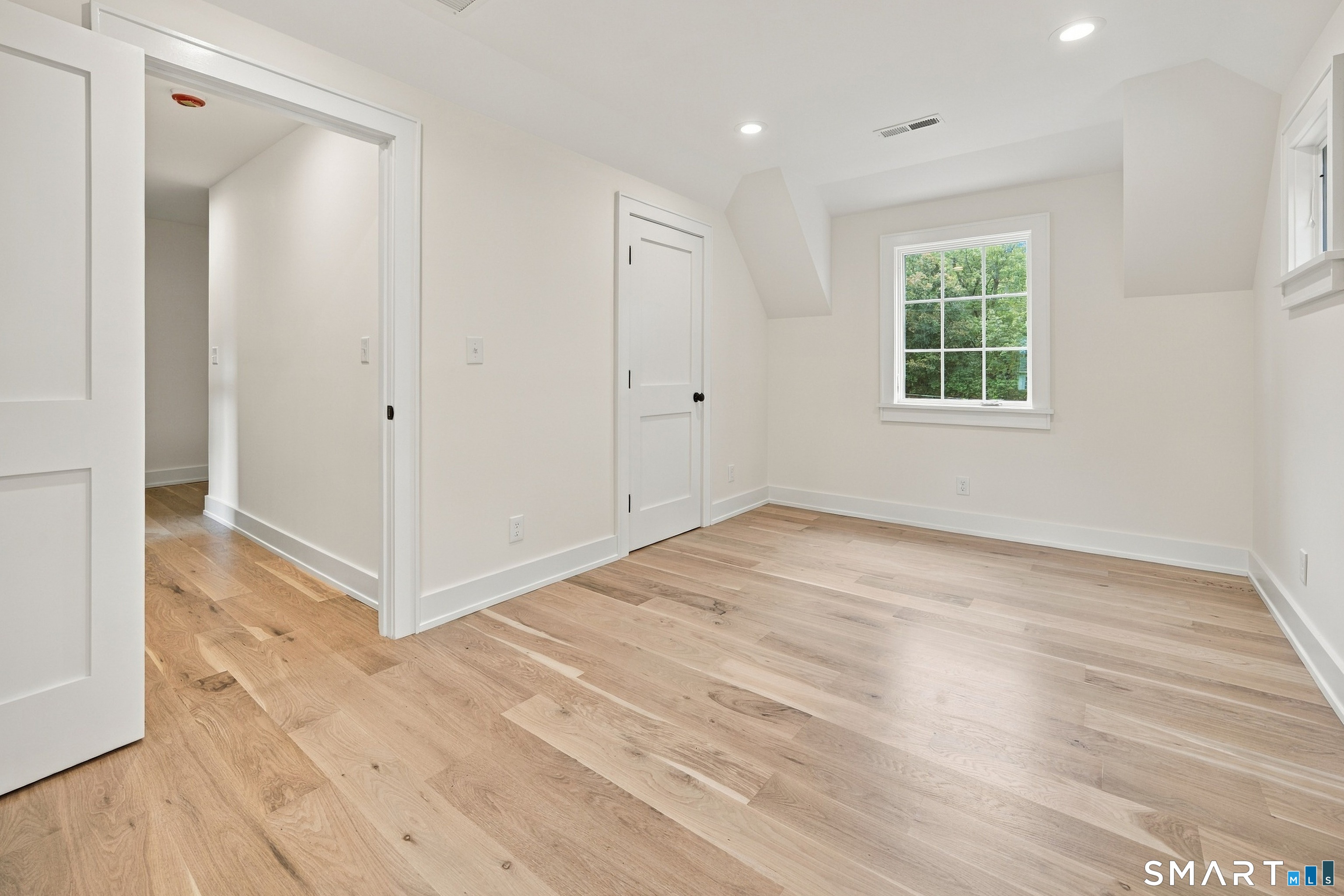 Empty room with light wood floors, white paneled doors, and a window overlooking trees.