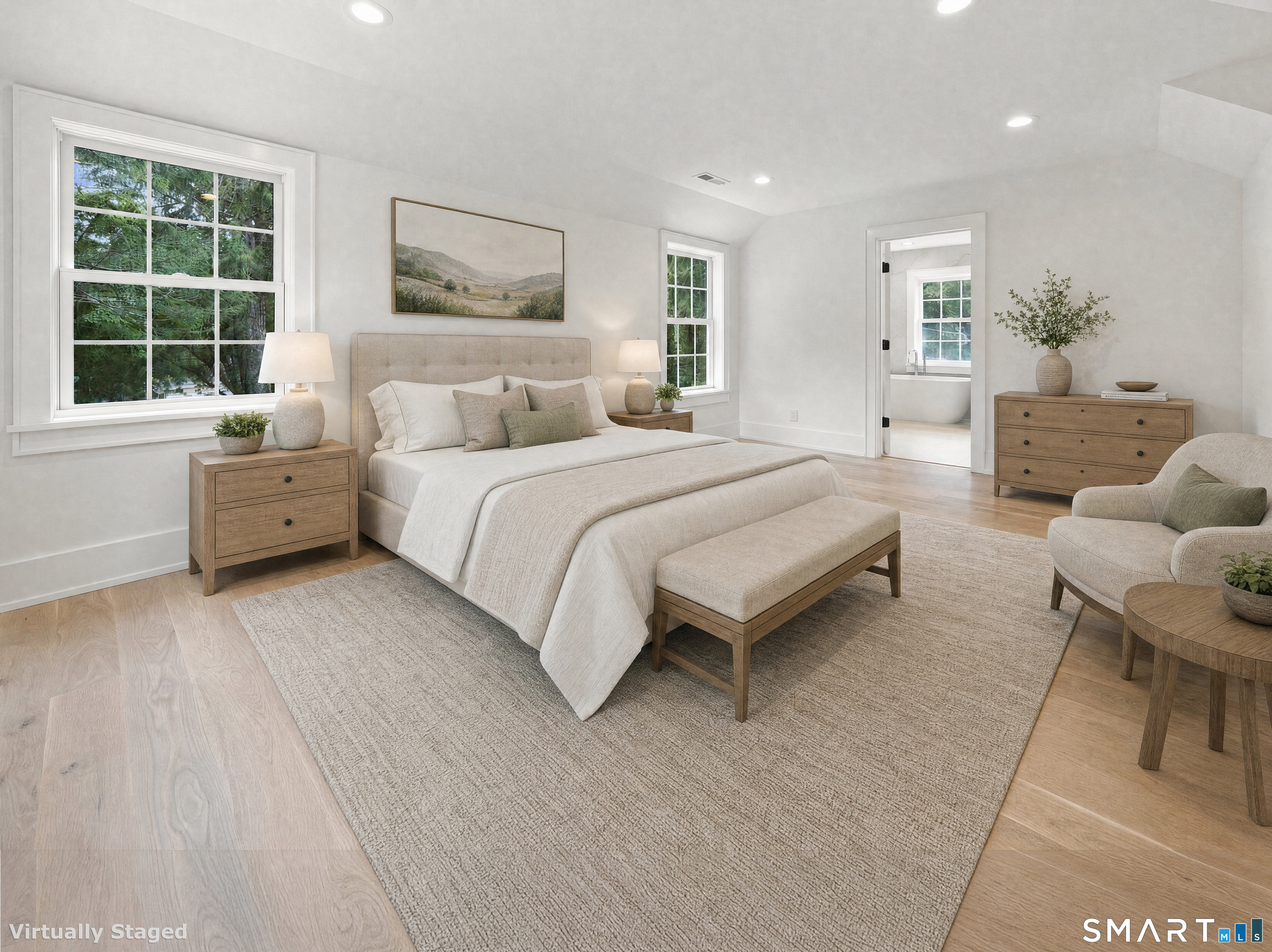 Staged bedroom with a large upholstered bed, light wood furniture, and windows overlooking trees.
