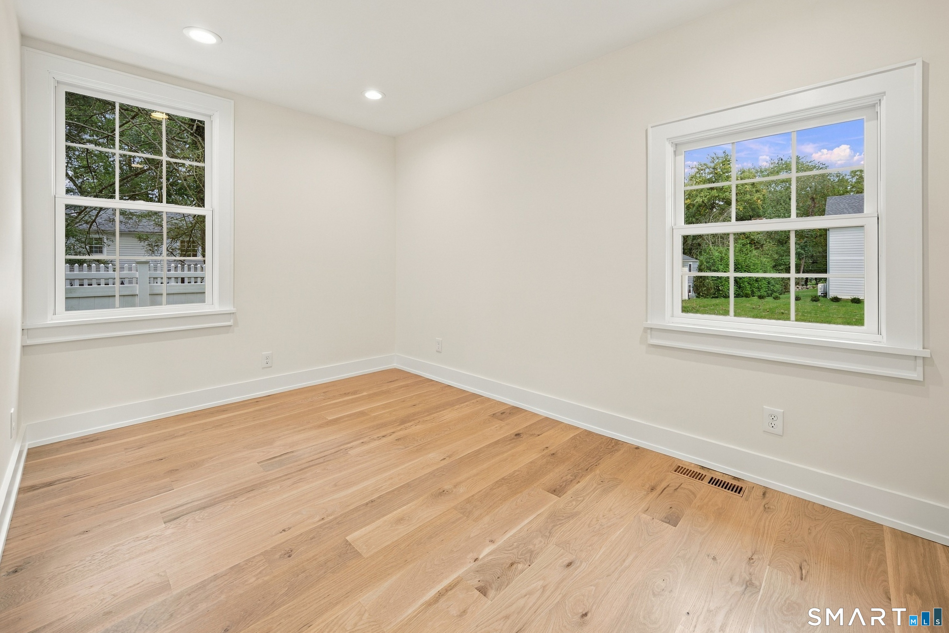 Empty room with light wood flooring and two large windows overlooking greenery.
