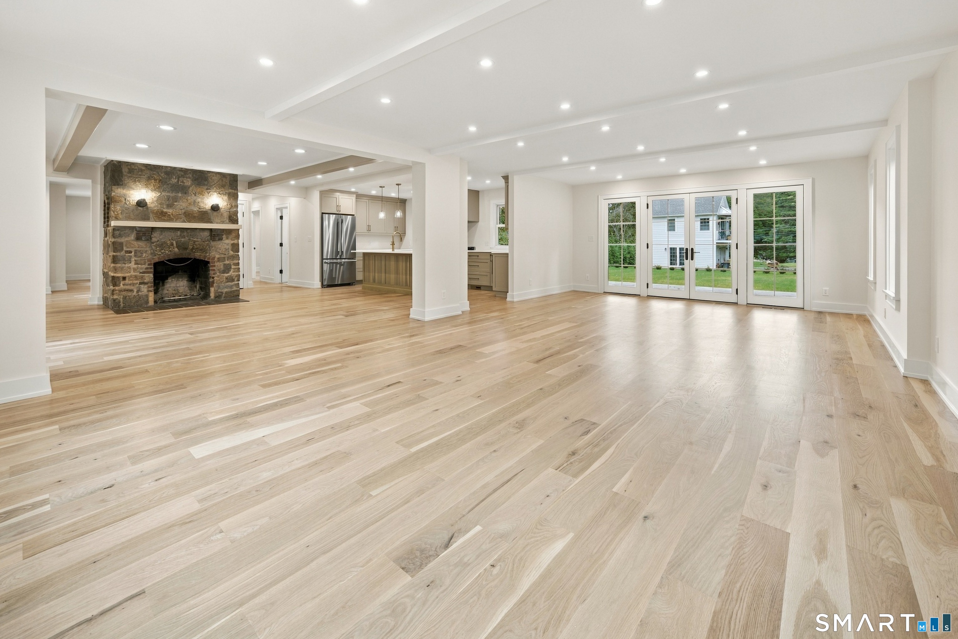 Unfurnished open living area with stone fireplace, pale wood floors, kitchen, and glass doors.